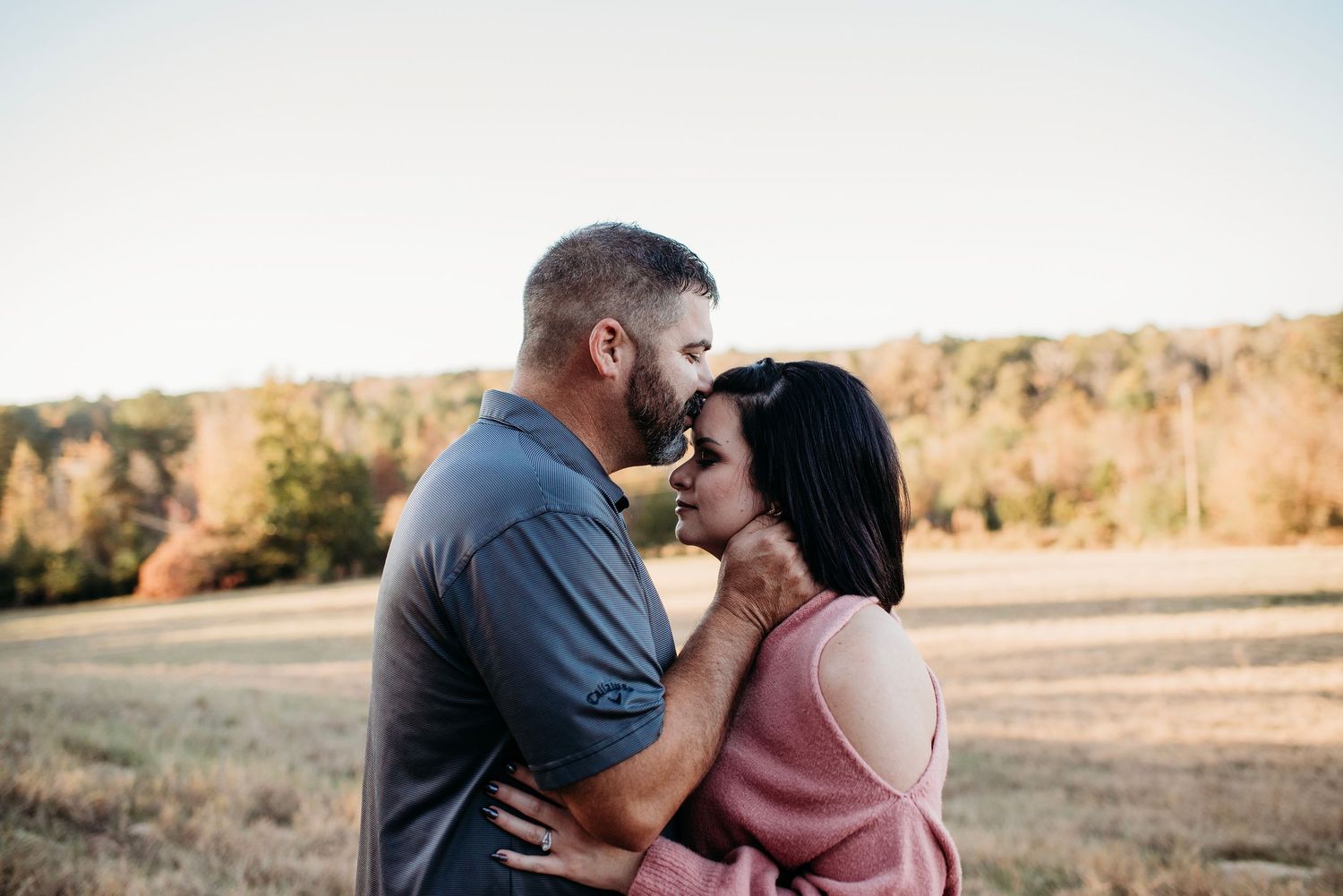 A couple shares an intimate moment together in a golden field during sunset with autumn trees in the background.