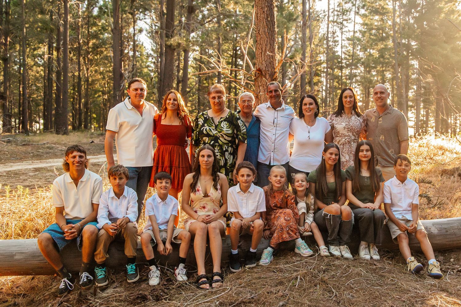 A large family group sits together on a fallen log in a warm, sunlit forest during golden hour.