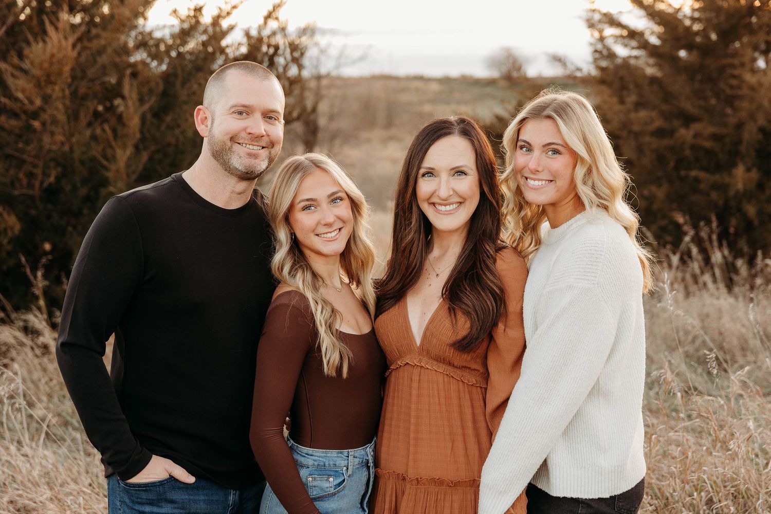 A group of four people pose together outdoors during golden hour with evergreen trees in the background.