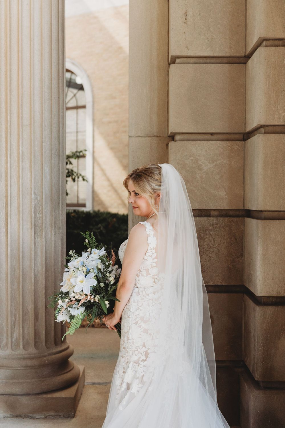 A bride in a lace wedding gown poses elegantly between classic stone columns in an architectural setting.