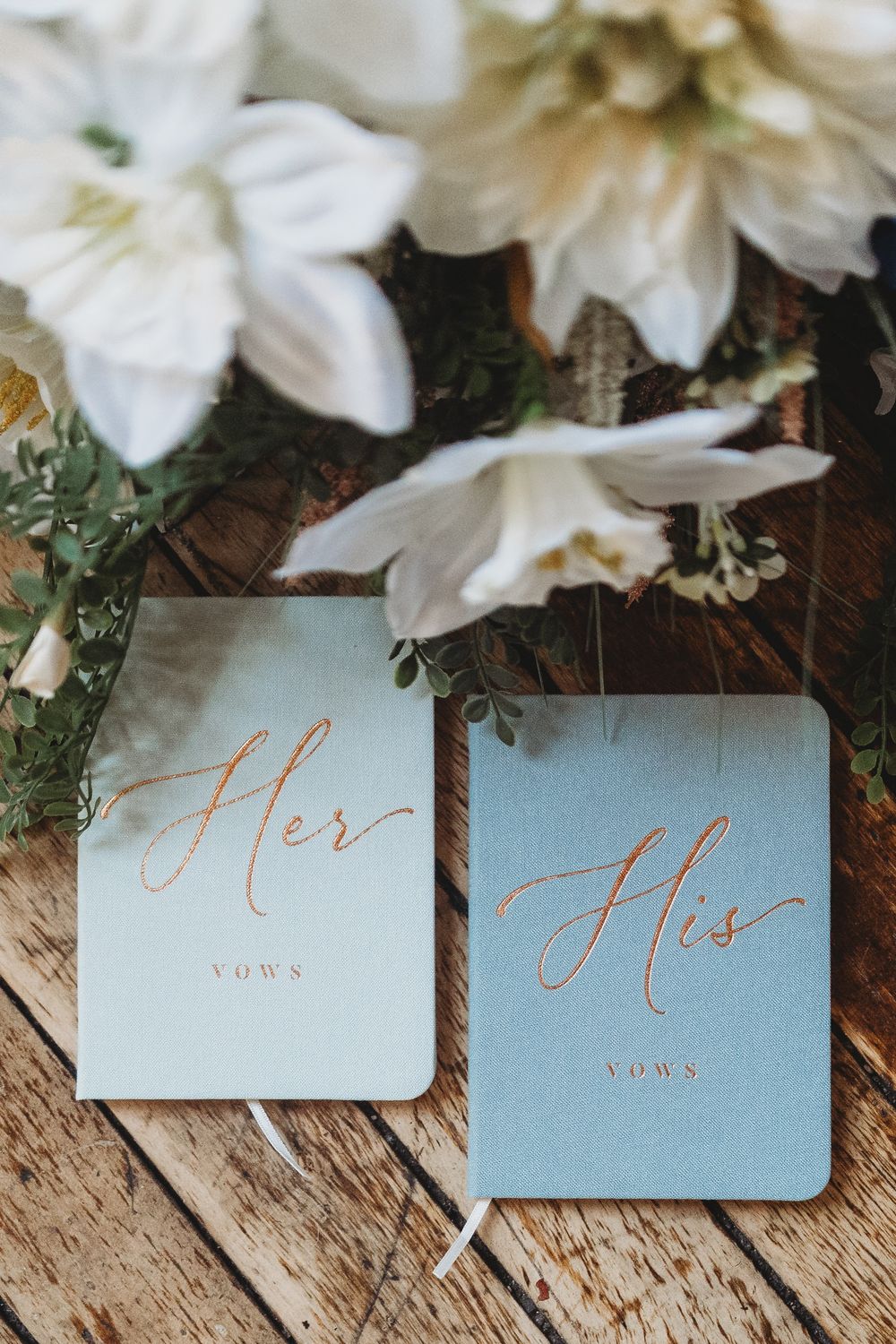 Wedding vows cards in light blue with rose gold lettering arranged on rustic wooden surface with white flowers.
