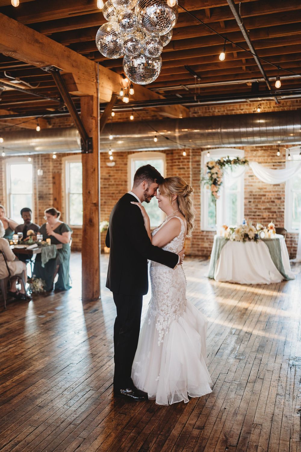 Wedding couple dances in rustic venue with exposed brick walls, wooden floors and disco ball lighting.