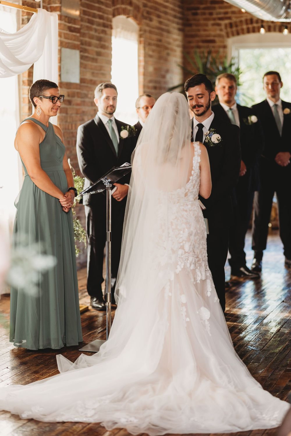 Wedding ceremony in a rustic brick-walled venue with sage green bridesmaids dresses and elegant white gown.