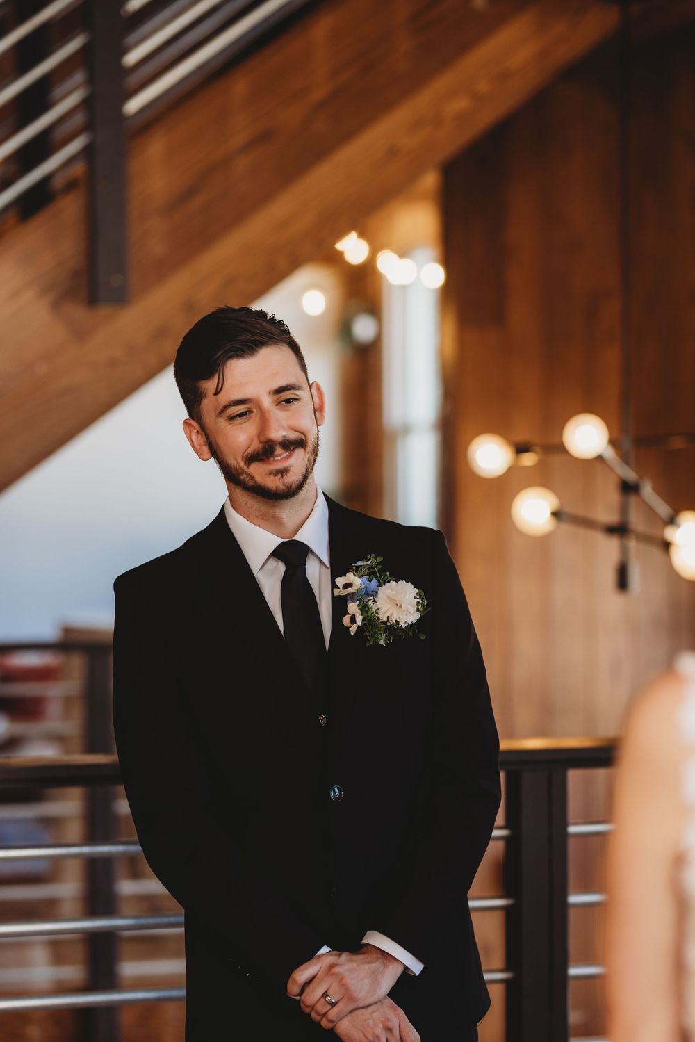 A groom in a black suit smiles during an emotional wedding ceremony moment.