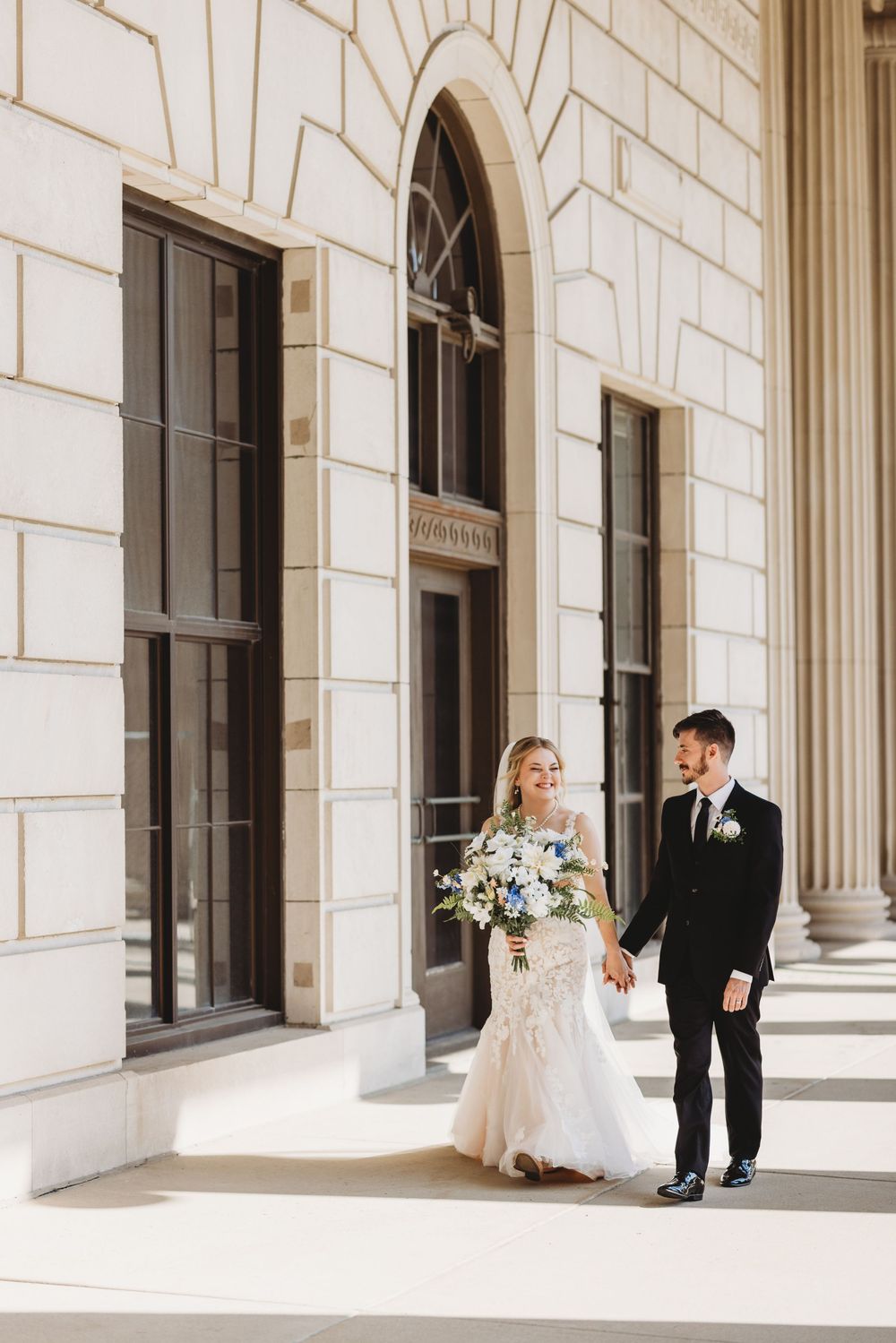 A couple in wedding attire poses elegantly along a white columned building with classic architecture.