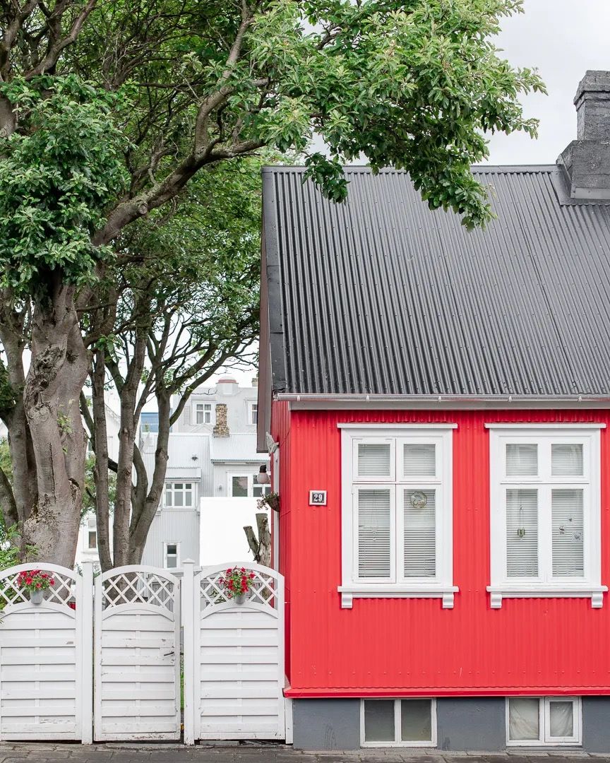 Red house with a grey roof behind a white fence and trees.
