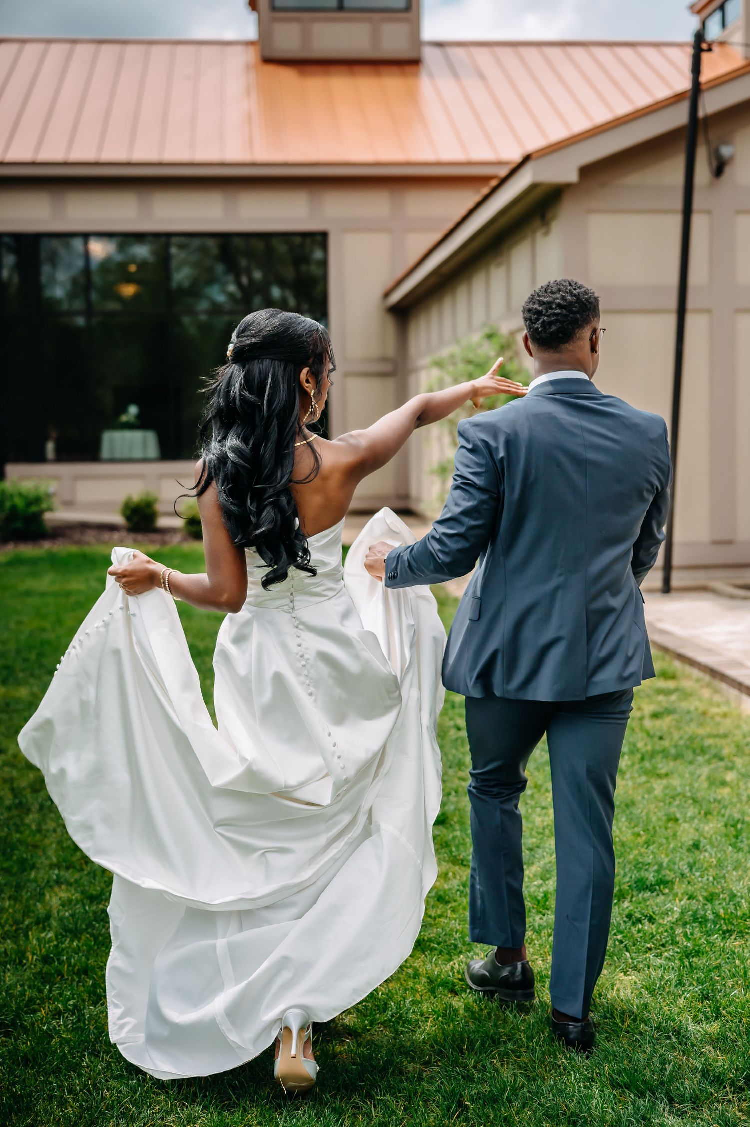 A couple in wedding attire playfully dances and twirls on a grassy lawn outside a building.