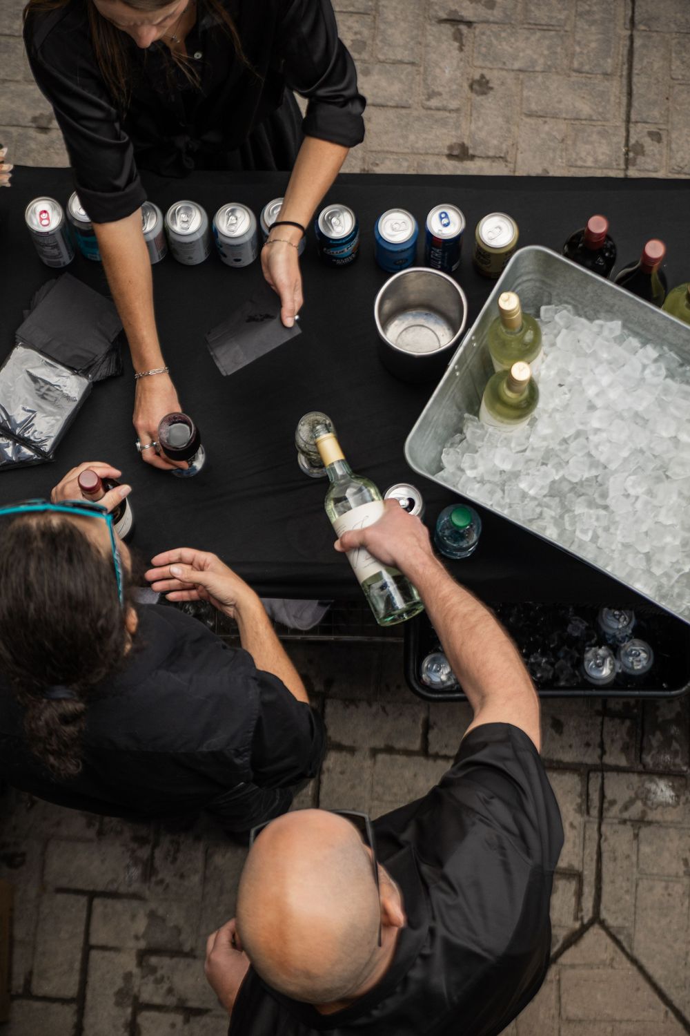Overhead view of people in black attire gathered around a bar with drinks and an ice bucket on a dark surface.