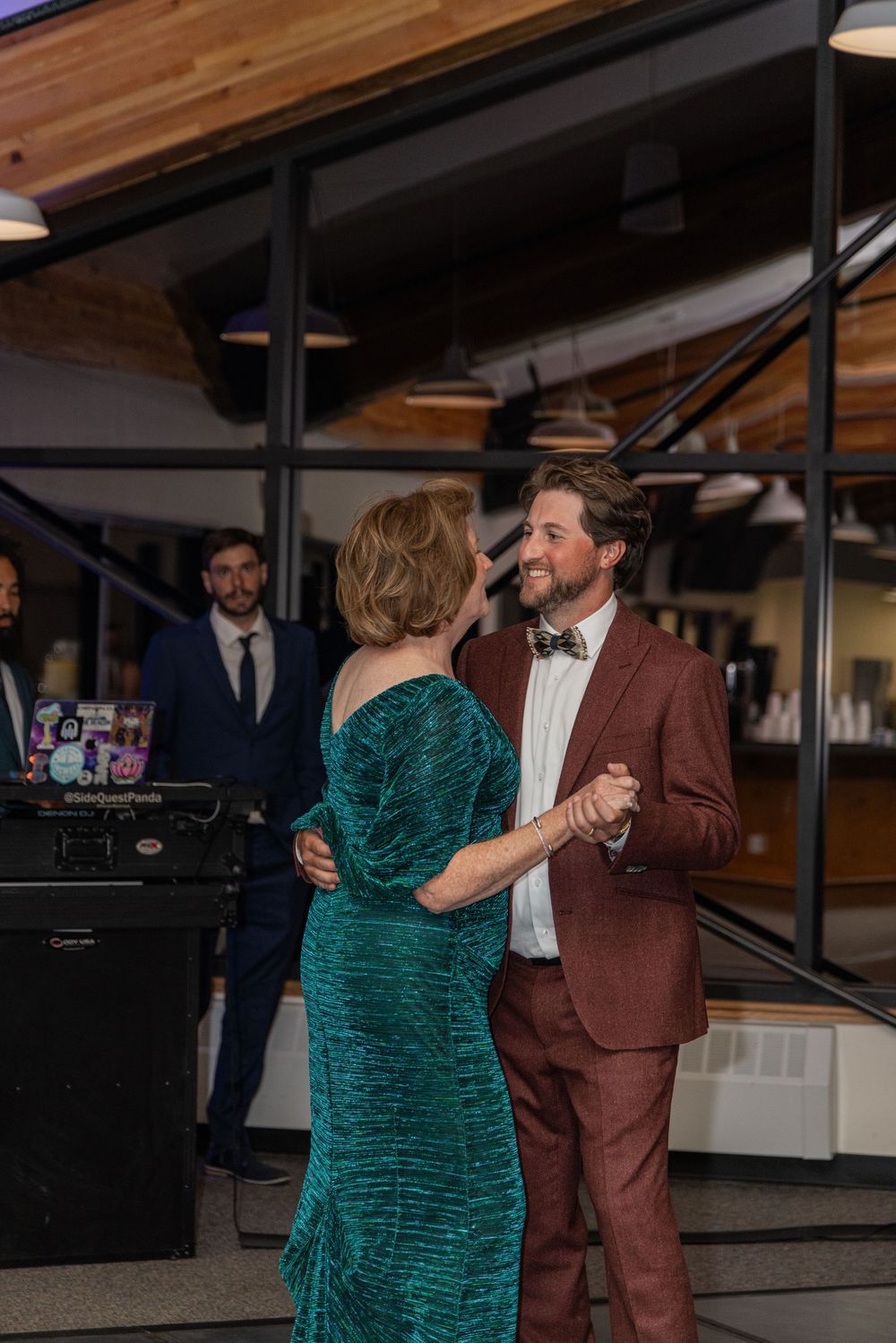 A couple dances together at an indoor venue, with the person wearing an elegant emerald green dress and brown suit.