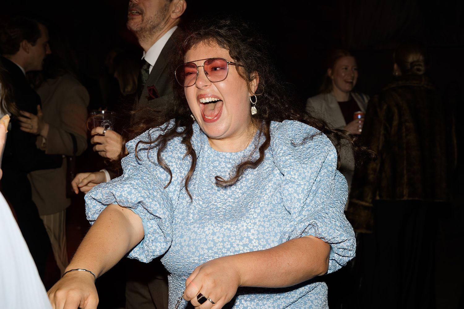 A person in a light blue dress with puff sleeves laughs joyfully while dancing at what appears to be a party or event.
