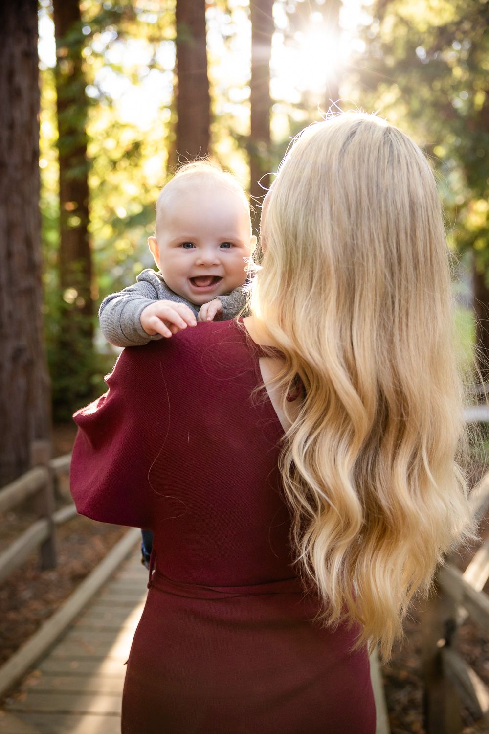 A mother and child share a playful moment on a wooden boardwalk in a sunlit redwood forest during golden hour in Los Altos.