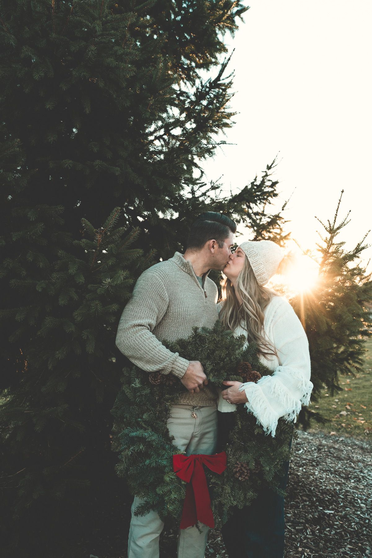A romantic couple shares a kiss while holding a Christmas wreath at sunset among evergreen trees at a park in Arlington Heights