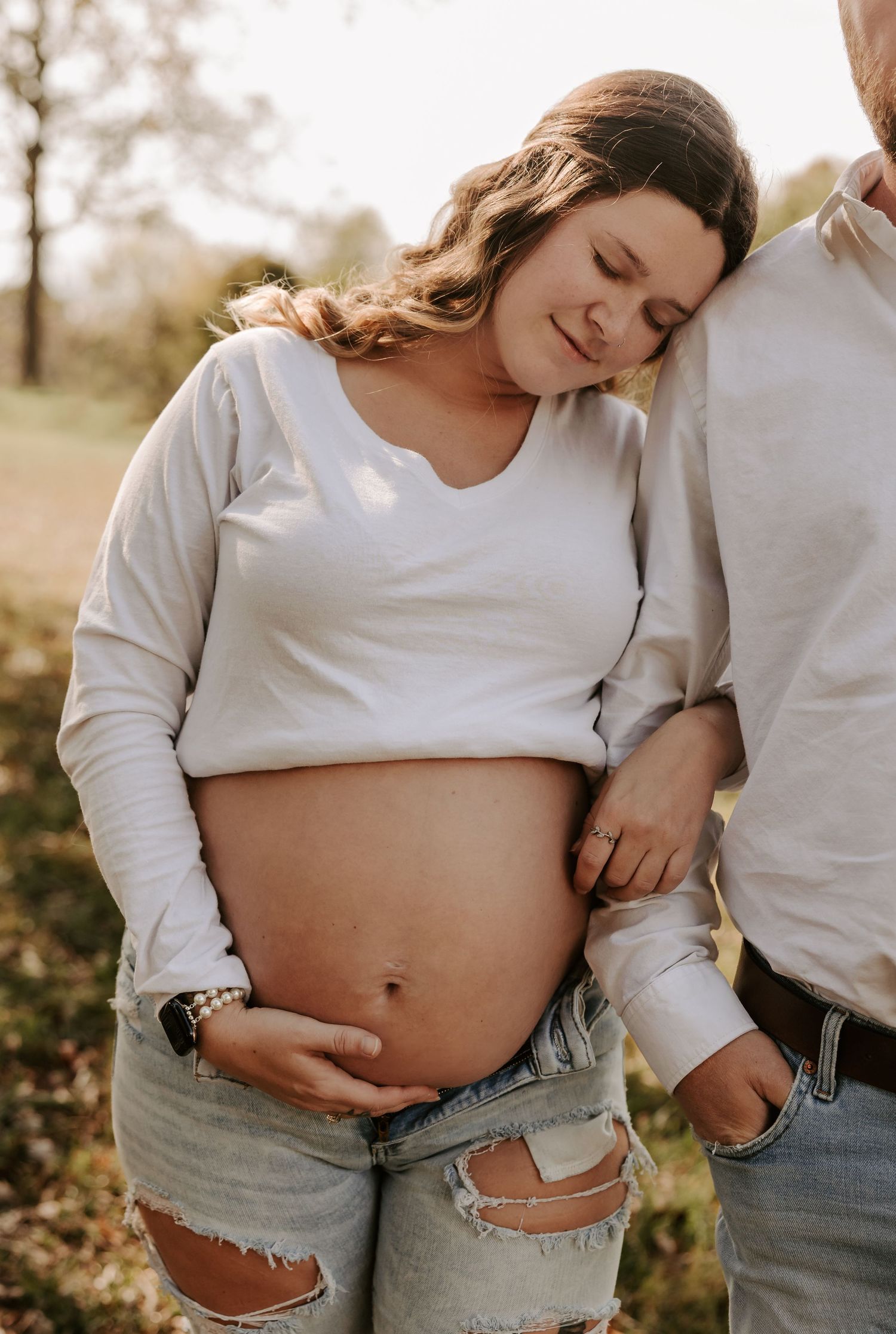 A candid maternity moment captured in warm sunlight with a natural outdoor background.