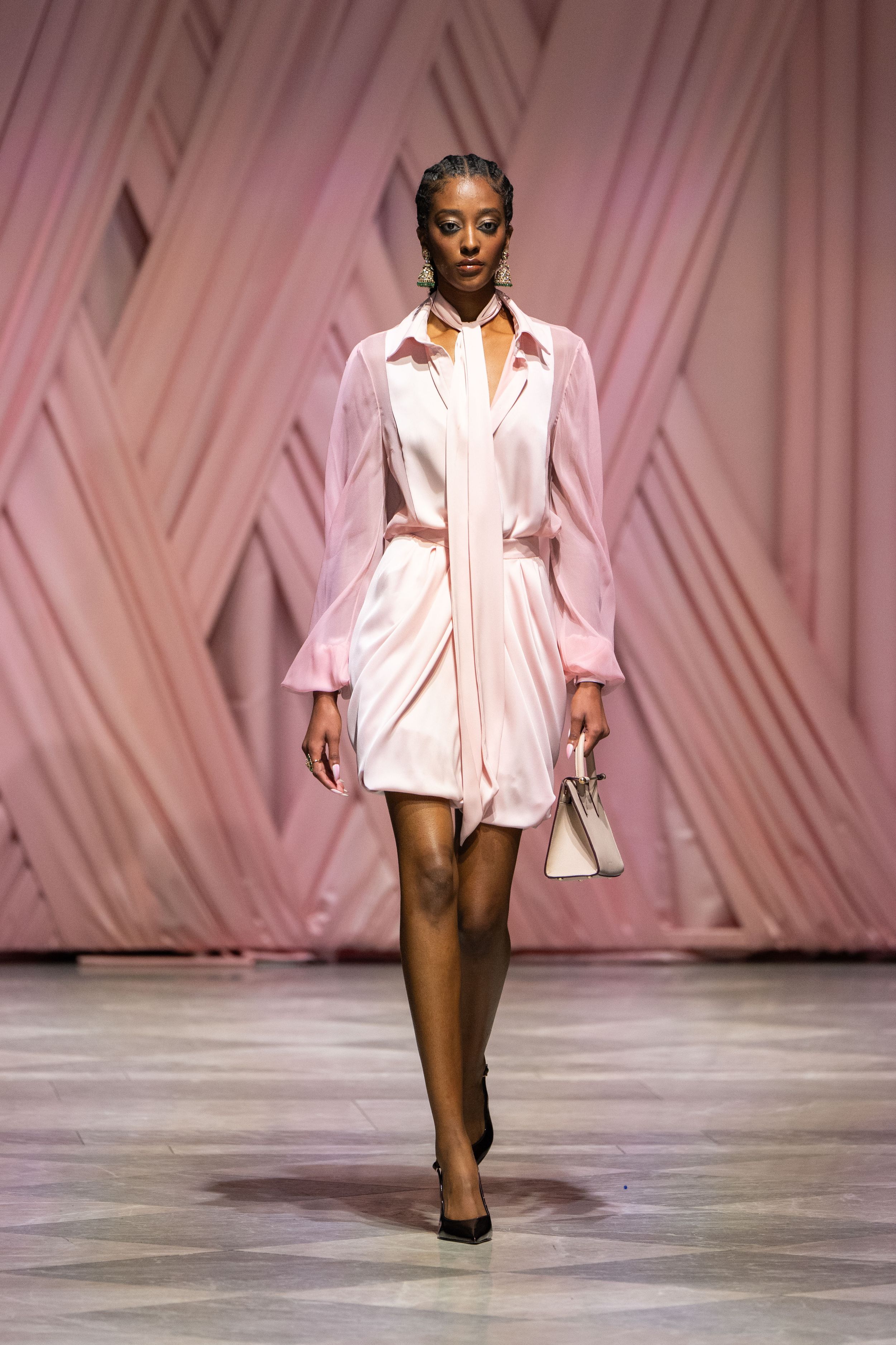 Fashion model walks runway wearing oversized white button-down shirt and heels against pink geometric backdrop.