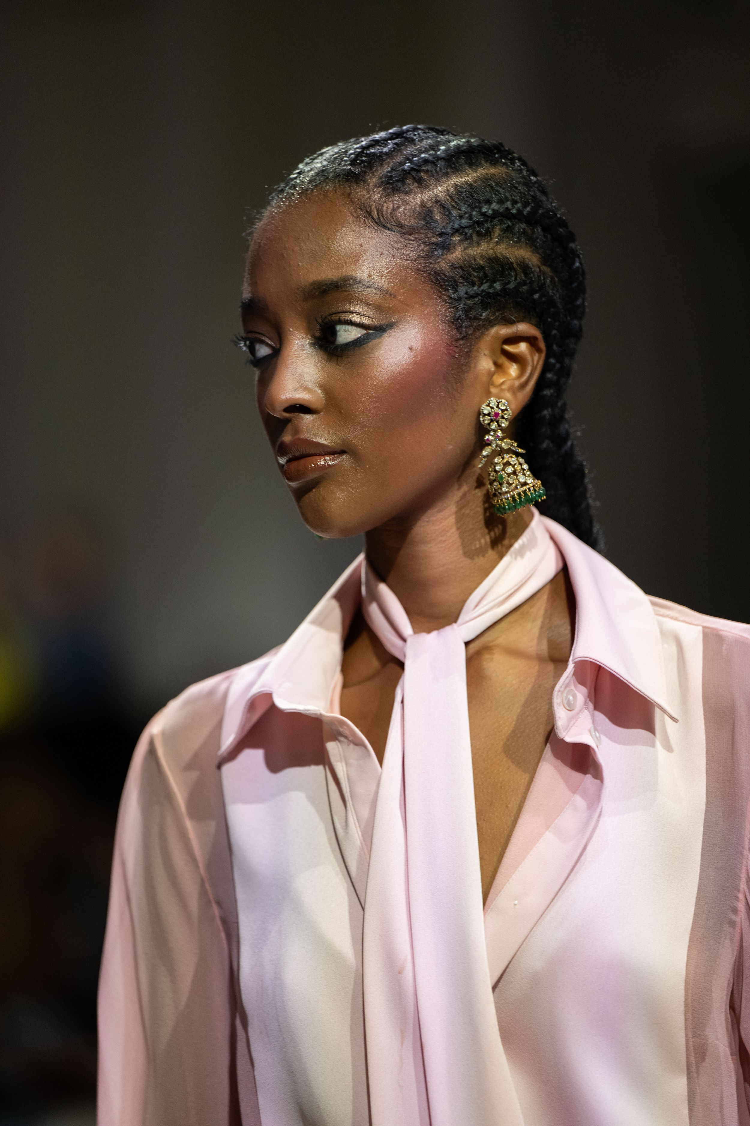 Model wearing pink blazer and cornrow braids walks runway at fashion show with dramatic side profile poses.