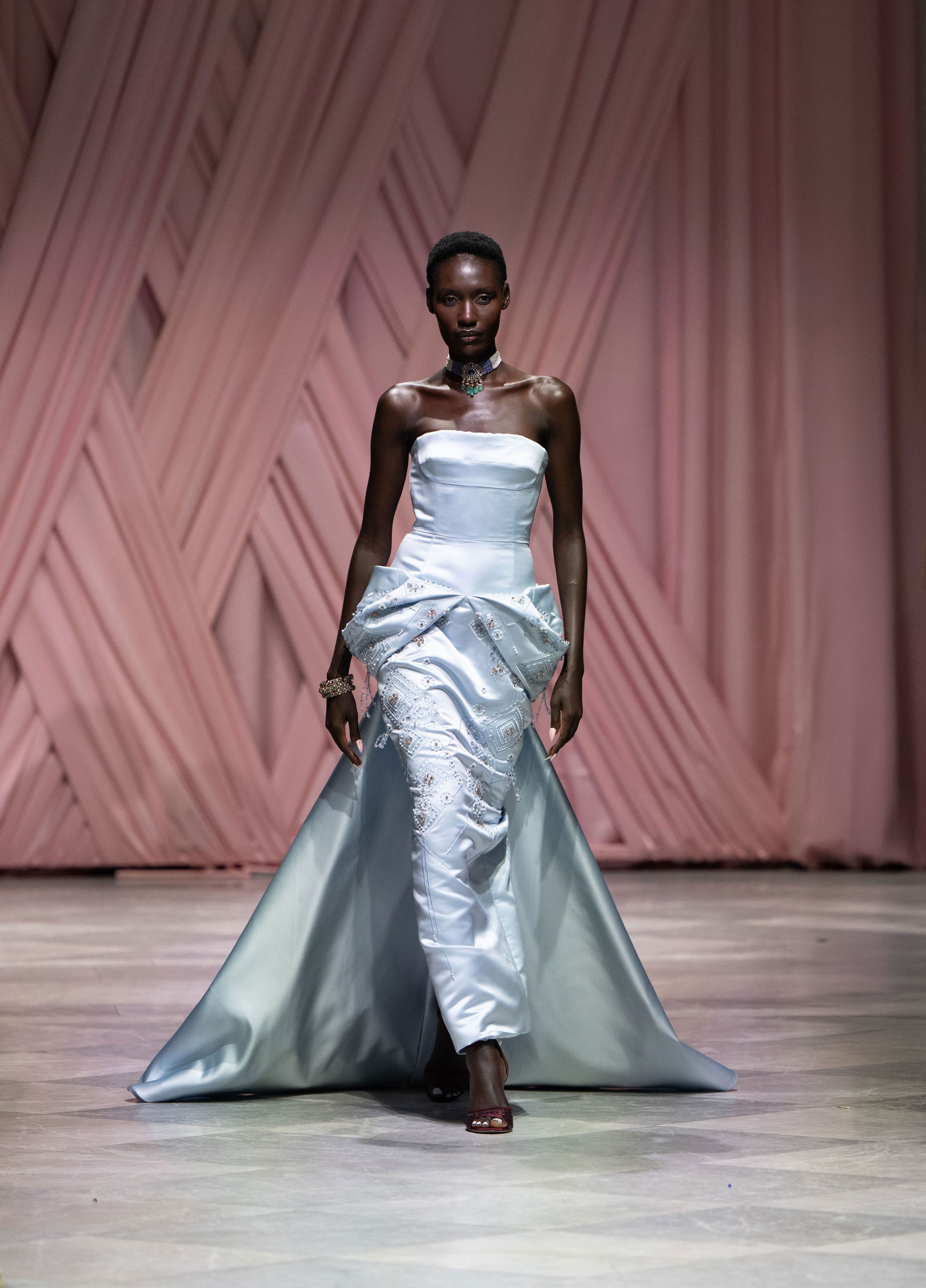 Fashion model walks runway in flowing metallic silver gown with dramatic train at haute couture fashion show.