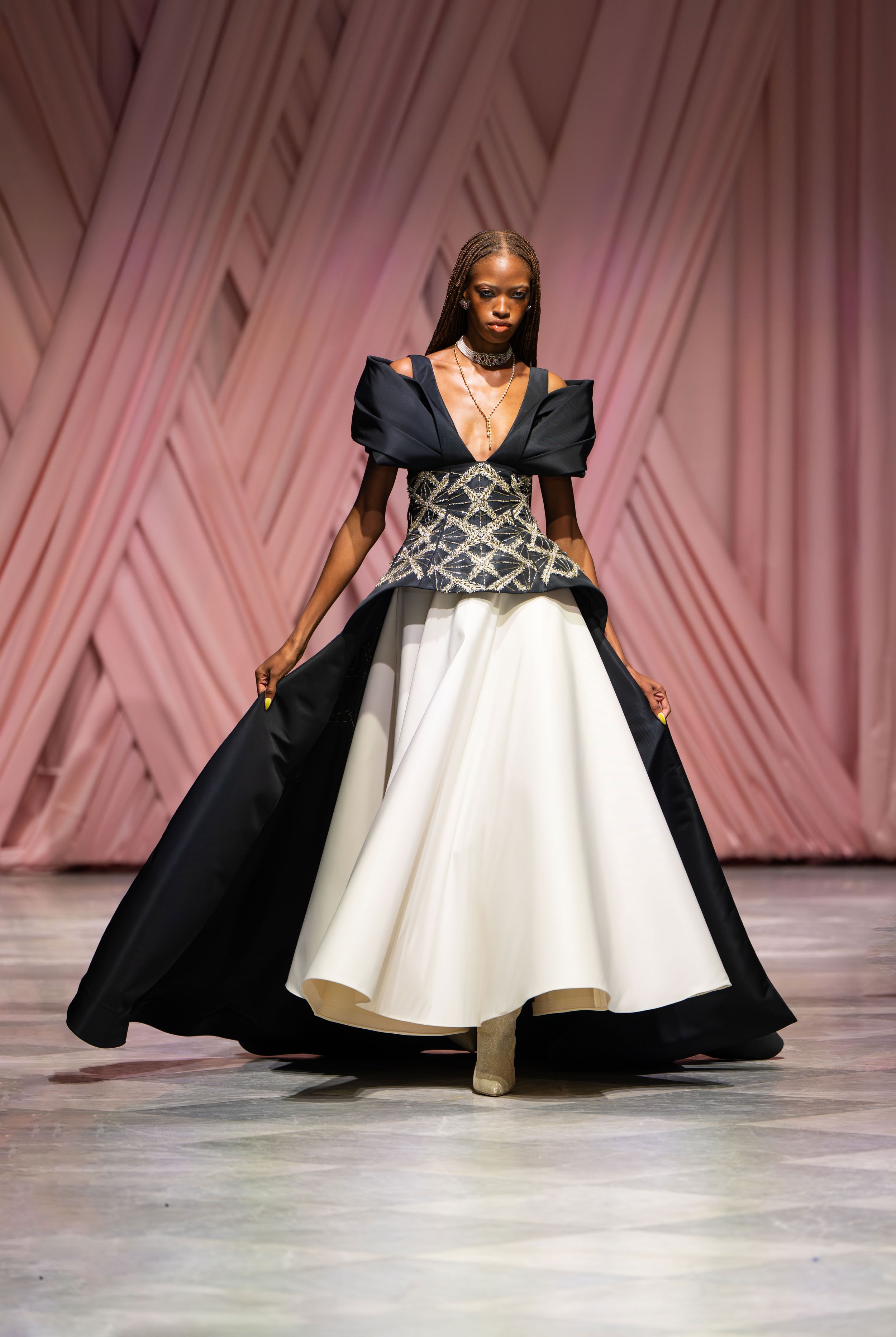 A dramatic black and white gown with exaggerated shoulders glides down the runway in a fashion show presentation.