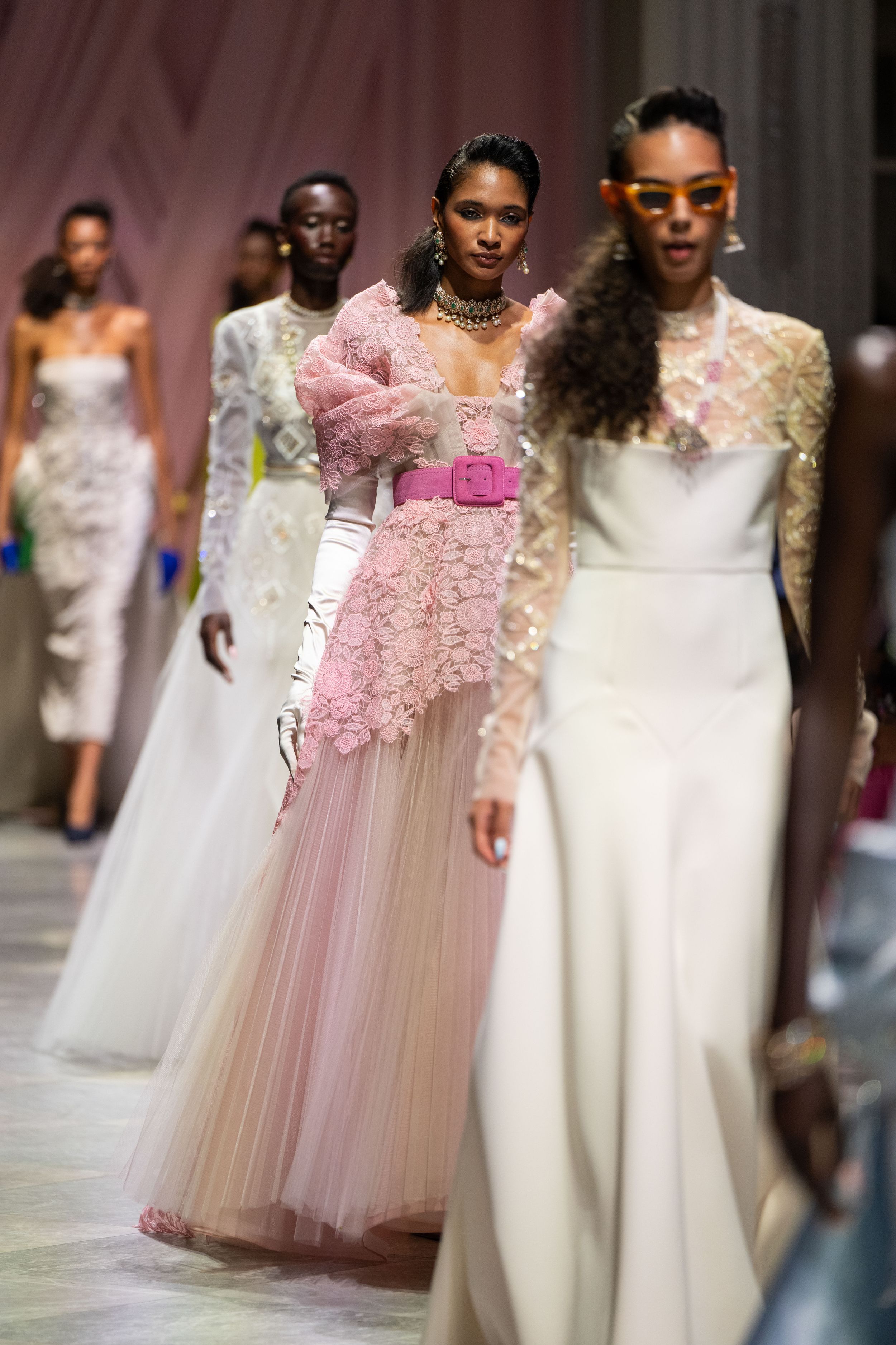 Models walk the runway in elegant white and pink wedding gowns during a bridal fashion show.