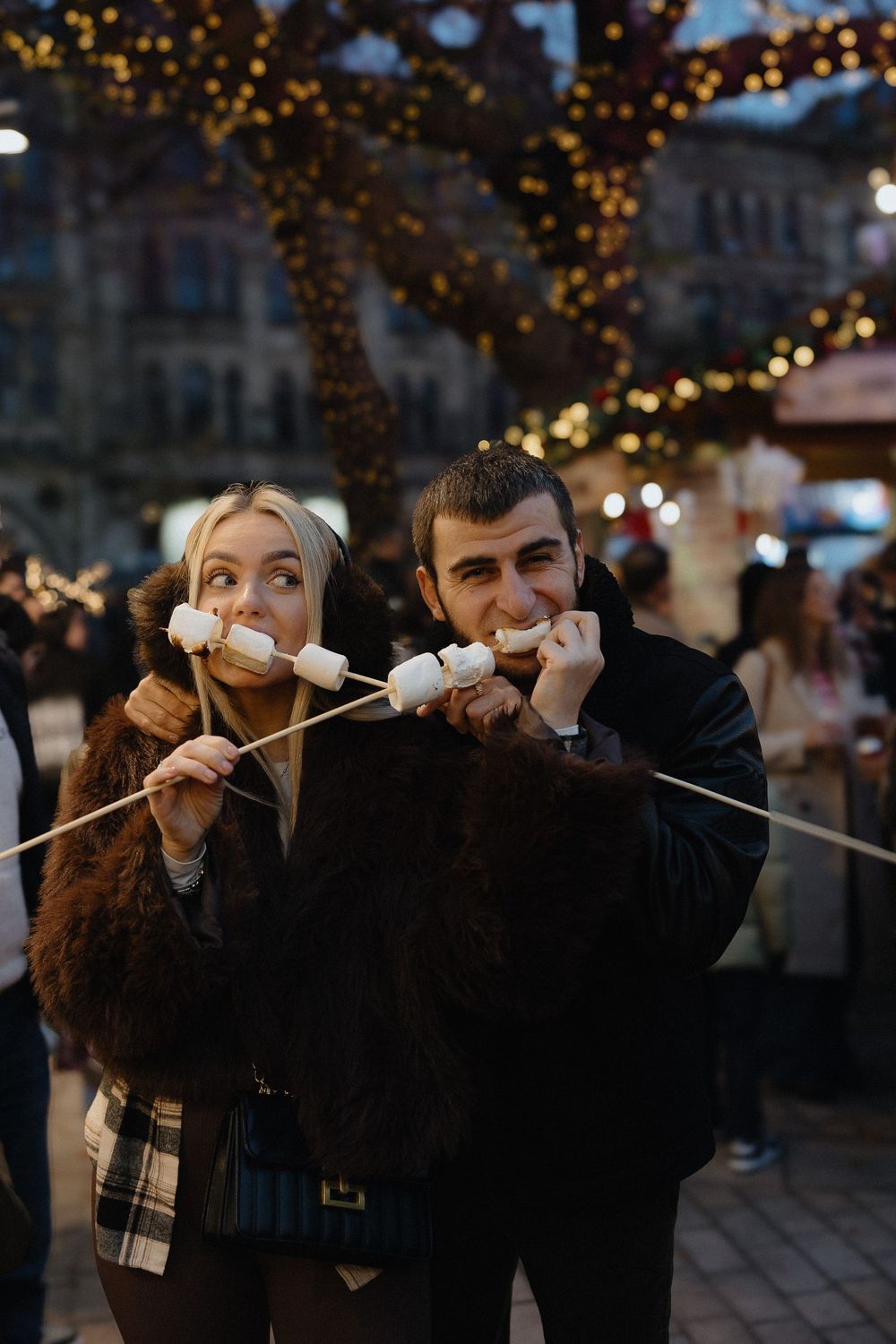 Friends competing in a marshmallow eating game at a festive outdoor market with string lights overhead.