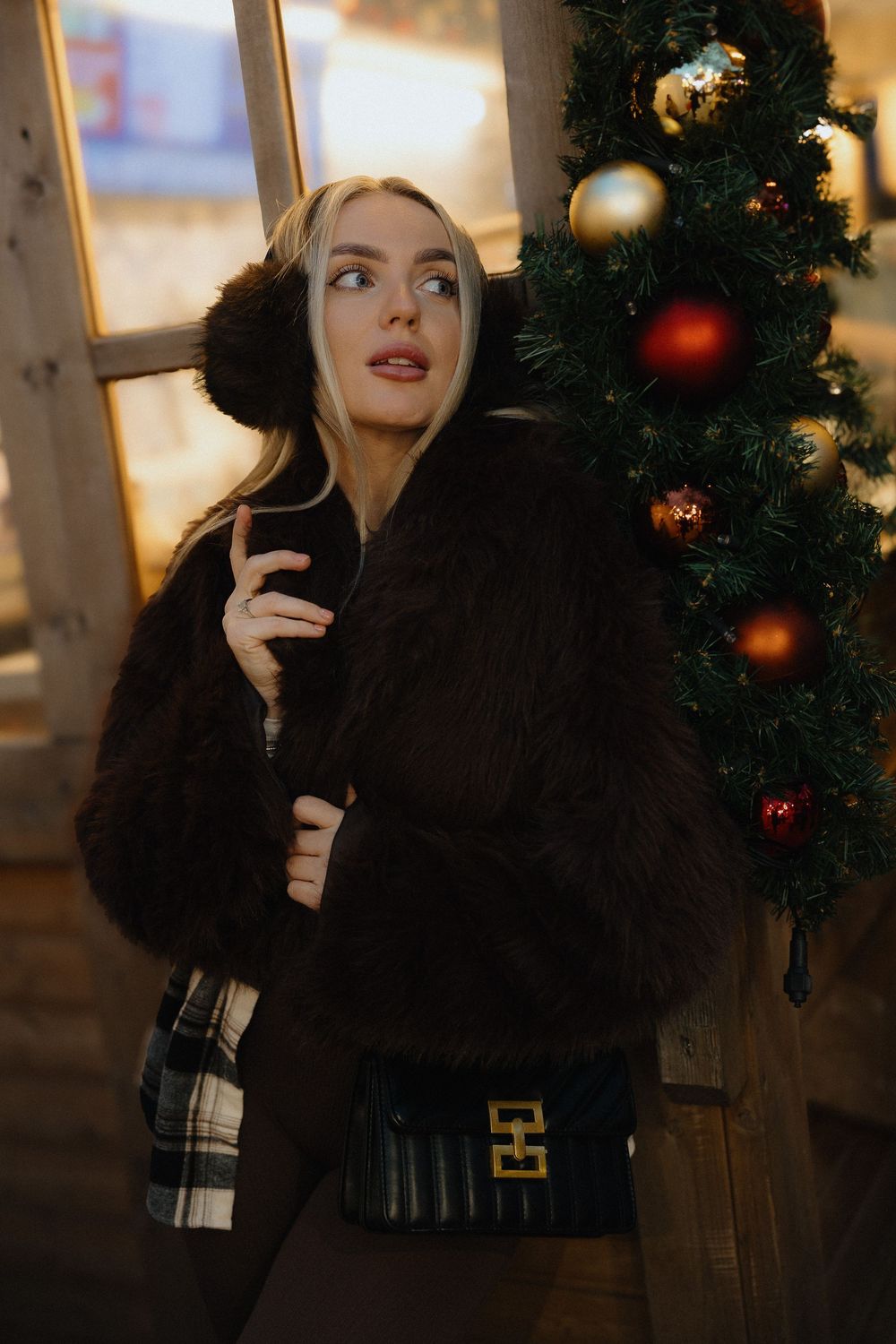 A person in a black winter coat poses against a backdrop of Christmas decorations and ornaments.