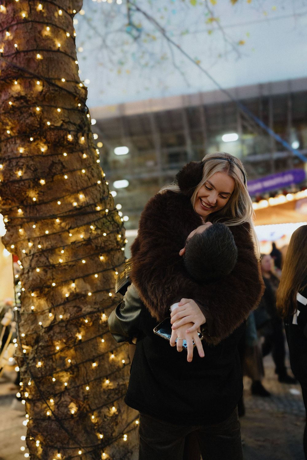 People embracing outdoors at night with a tree decorated in twinkling holiday lights in the background.