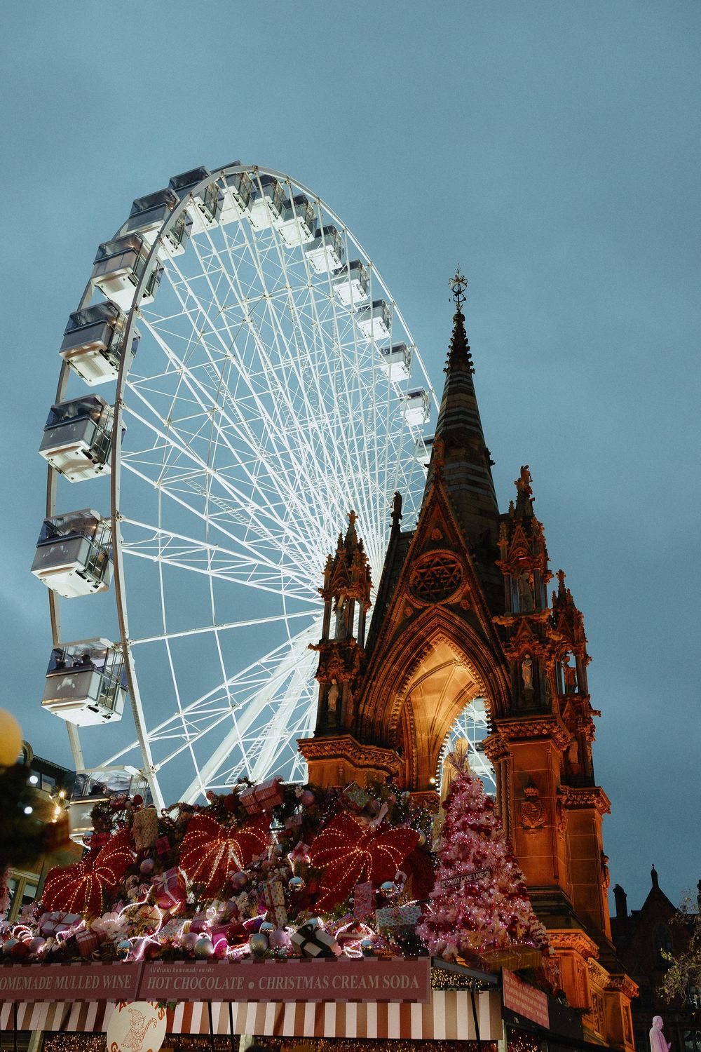 A white Ferris wheel illuminated at dusk stands beside a Gothic cathedral with pink cherry blossoms in the foreground.