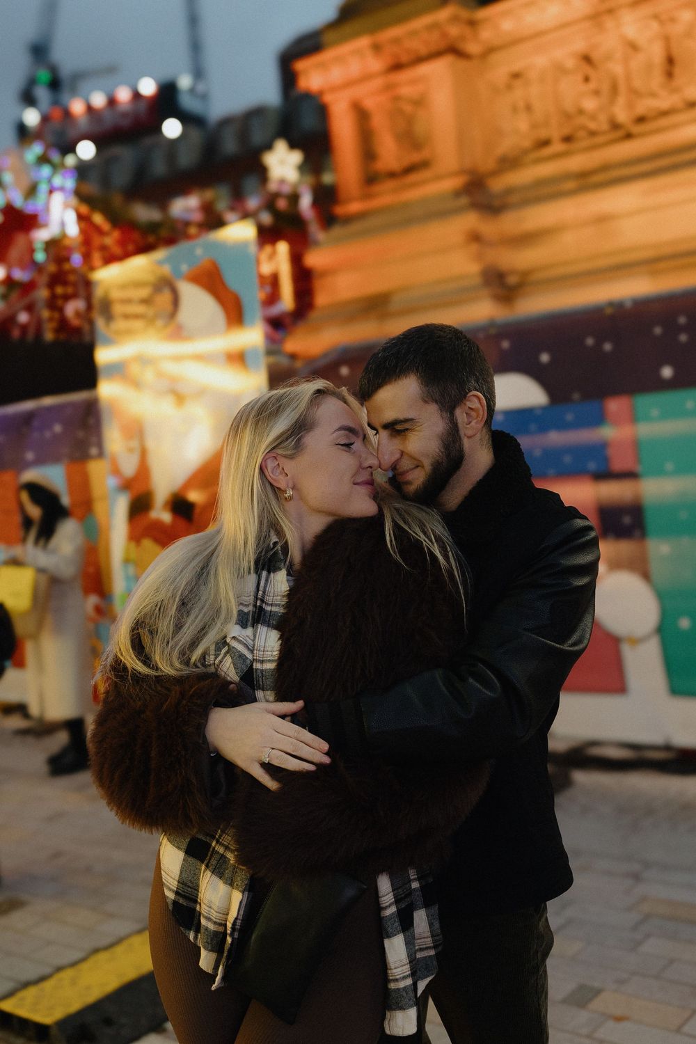 A romantic couple embraces and kisses at night with festive lights and buildings in the background.