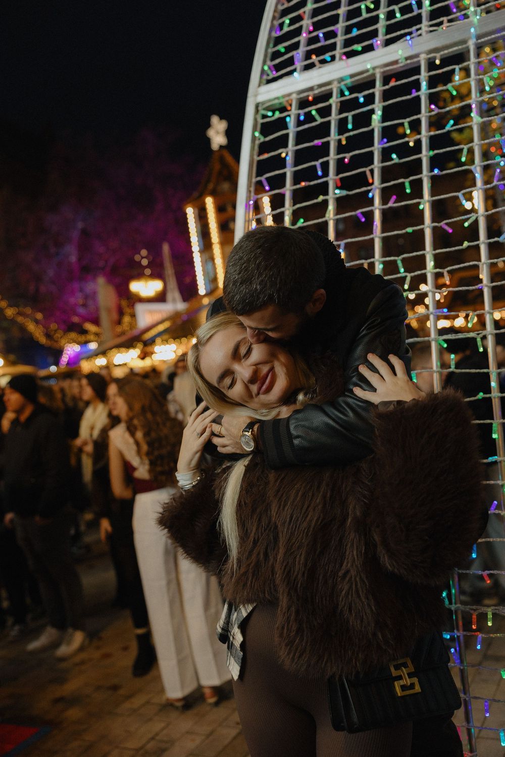 A couple embracing at night surrounded by colorful Christmas lights and holiday decorations.