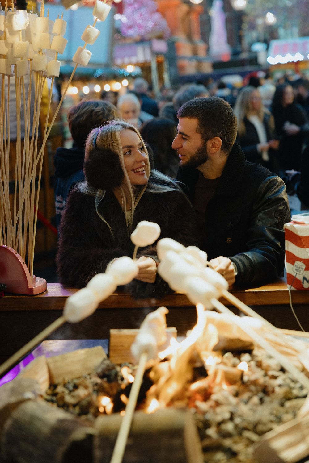 People gather around a table at an evening market or festival with glowing lights and wooden skewers in the foreground.