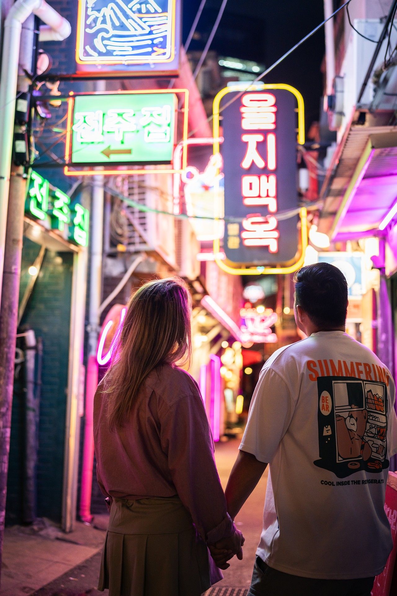 A couple holds hands while walking through a neon-lit alley in South Korea with glowing Korean signs at night.