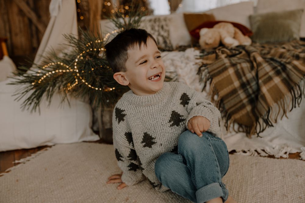 A laughing child sits on the floor wearing a star-patterned sweater with Christmas decorations in the background.