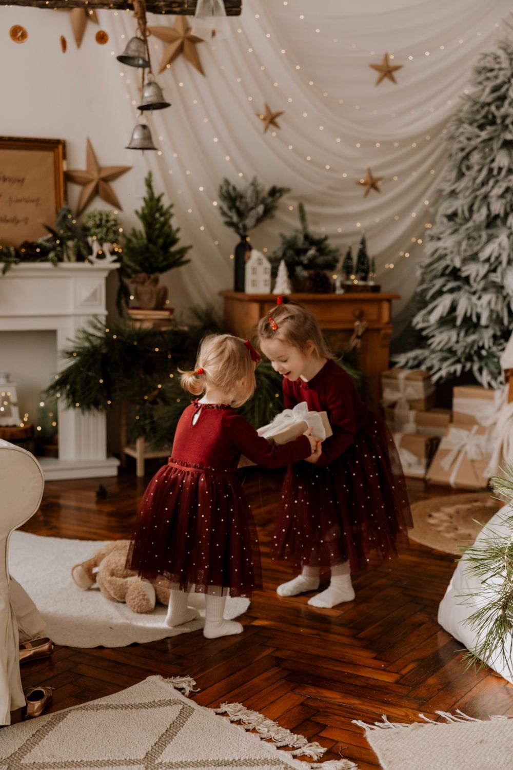 Festive Christmas scene with matching burgundy dresses against white drapes with gold stars and snowy decorations.