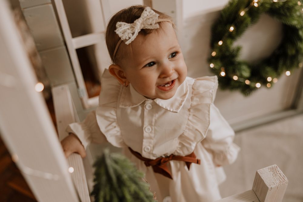 A baby in a white lace dress sits near twinkling Christmas lights and holiday decorations.
