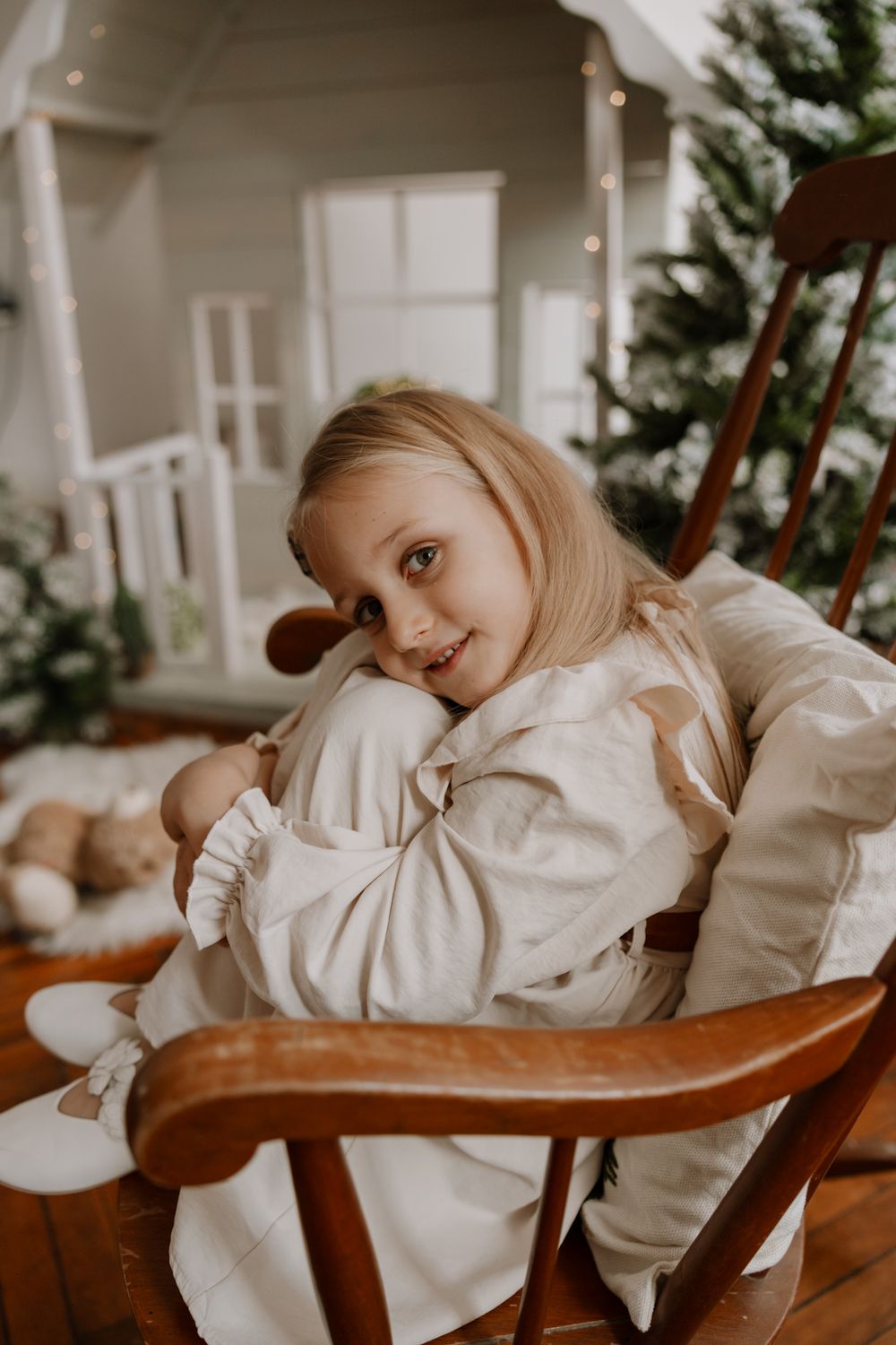 Child in white dress rests on wooden rocking chair near decorated Christmas tree in cozy indoor setting.
