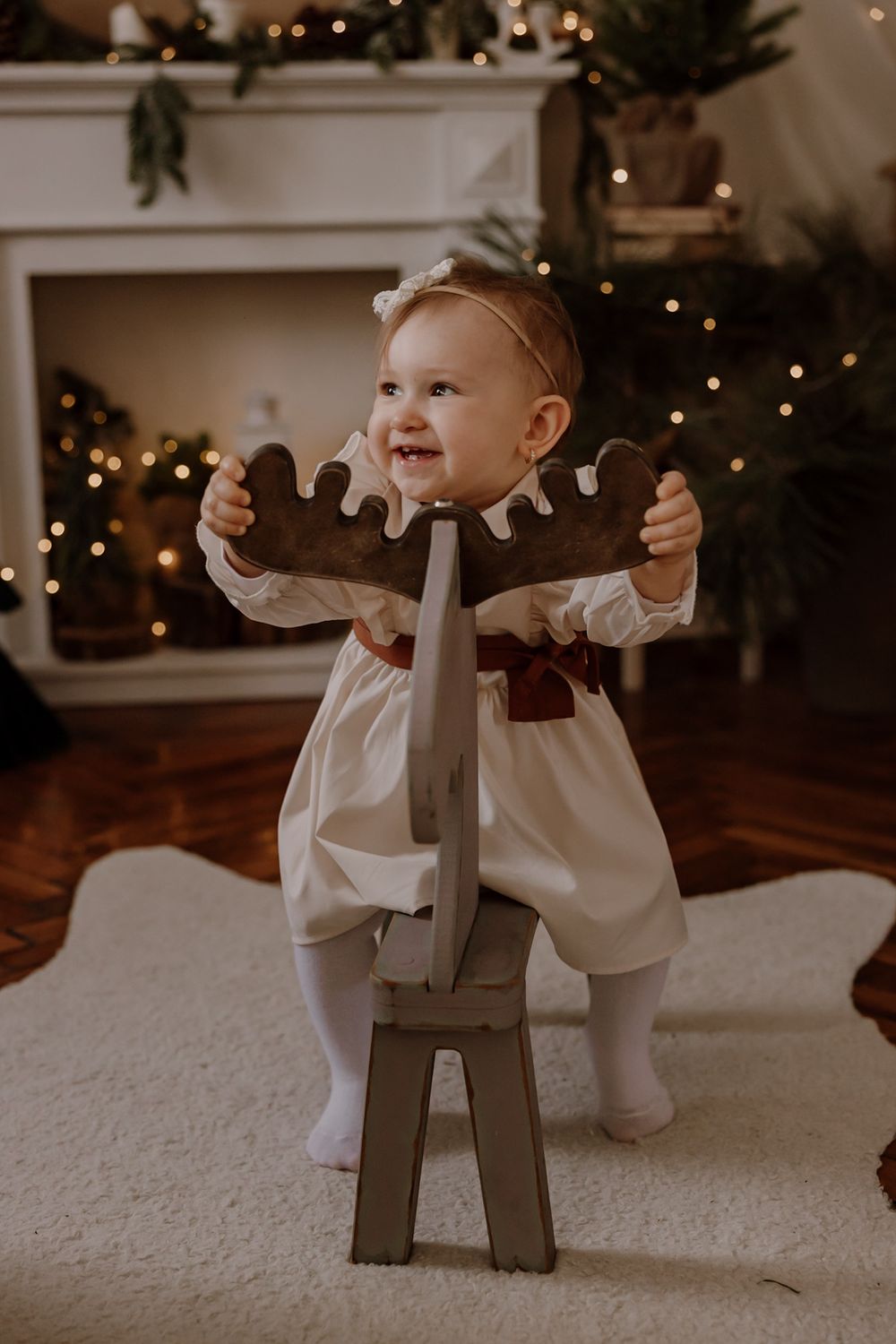 Small child in white clothing plays with a wooden reindeer decoration on a carpeted floor with Christmas lights in background.