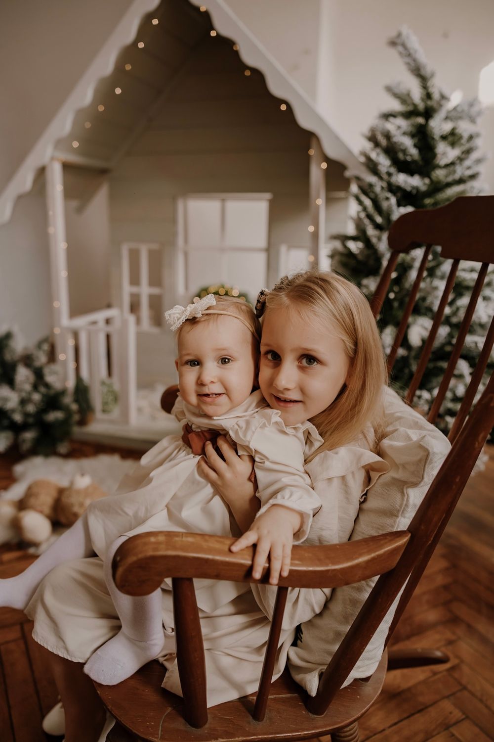 Two children in white outfits share sweet moments while sitting in a wooden rocking chair during a holiday photo session.