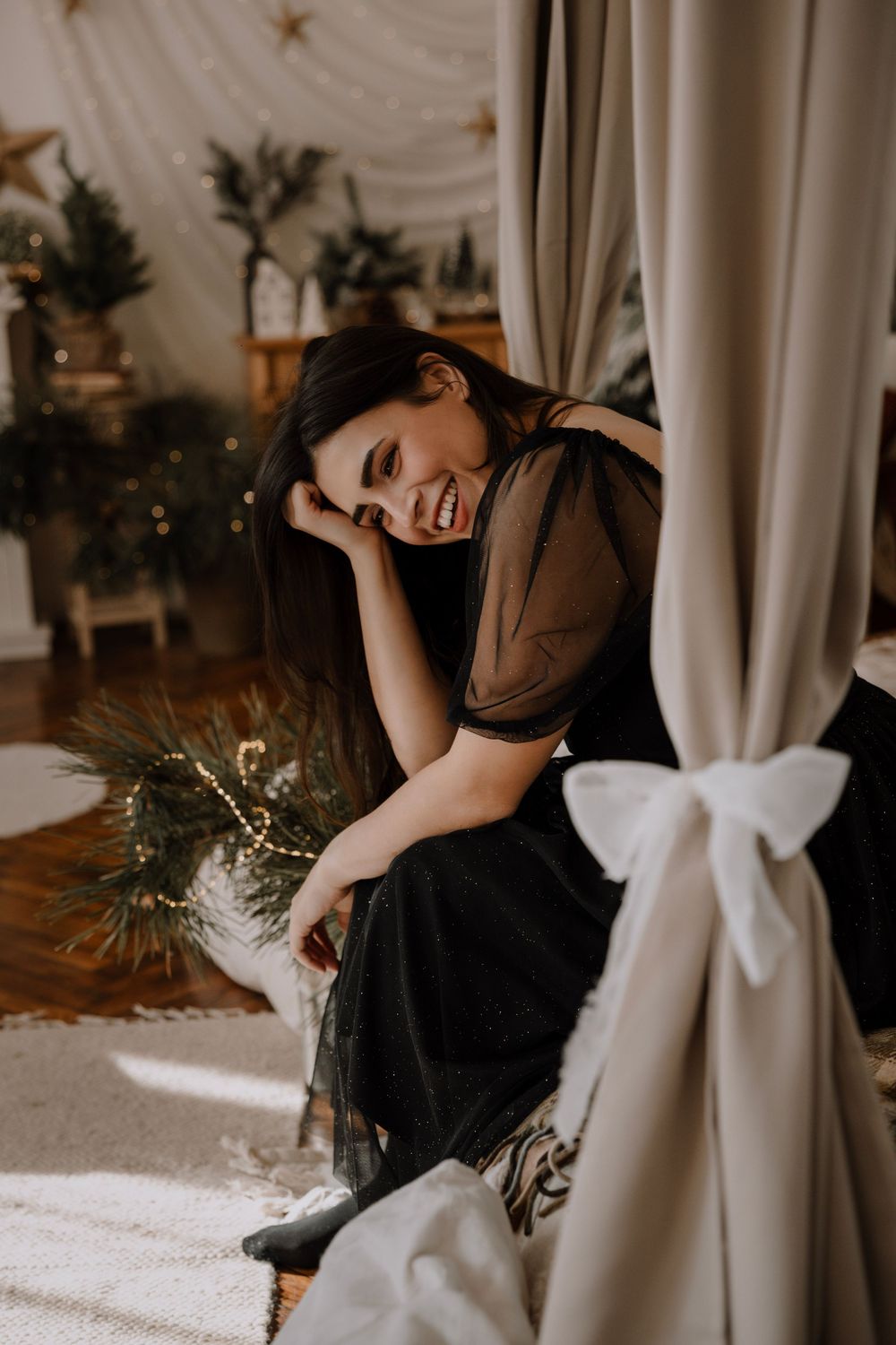 A person in black attire poses artistically near white curtains in a moody indoor setting.