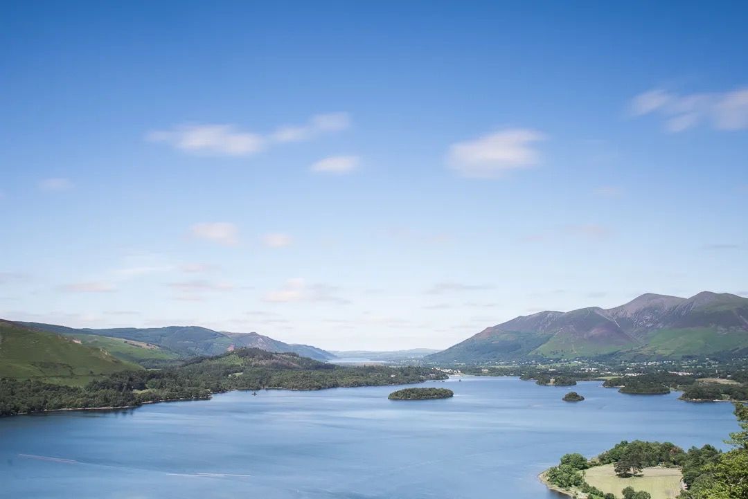 Wide view of a calm lake surrounded by rolling green hills under a blue sky.