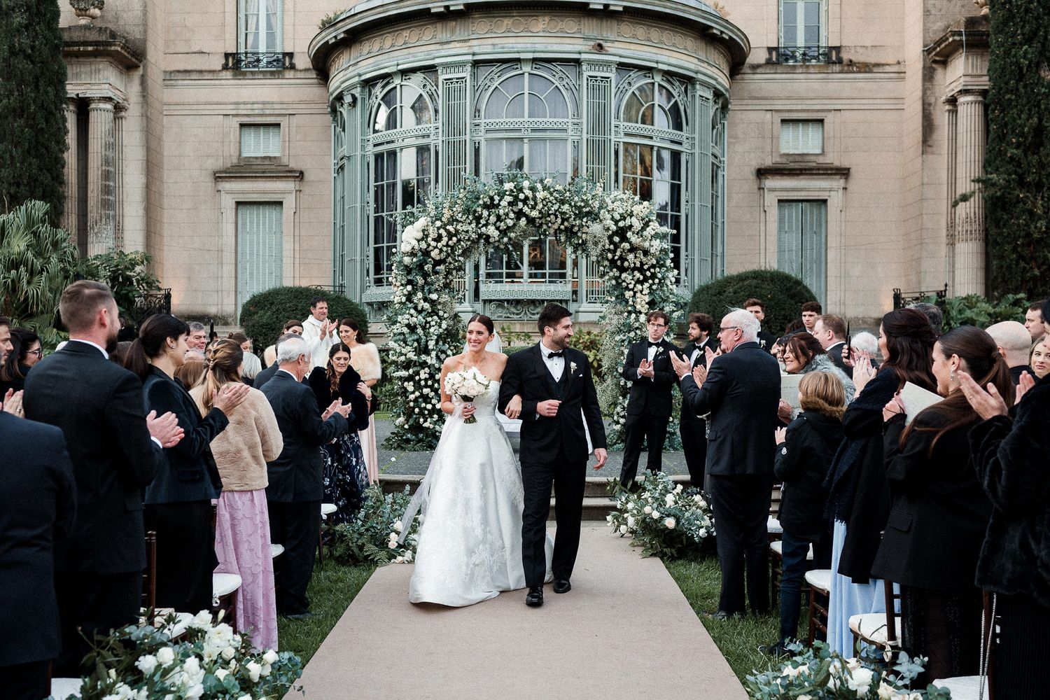 Newlyweds walking down flower-lined aisle while guests celebrate their exit.
