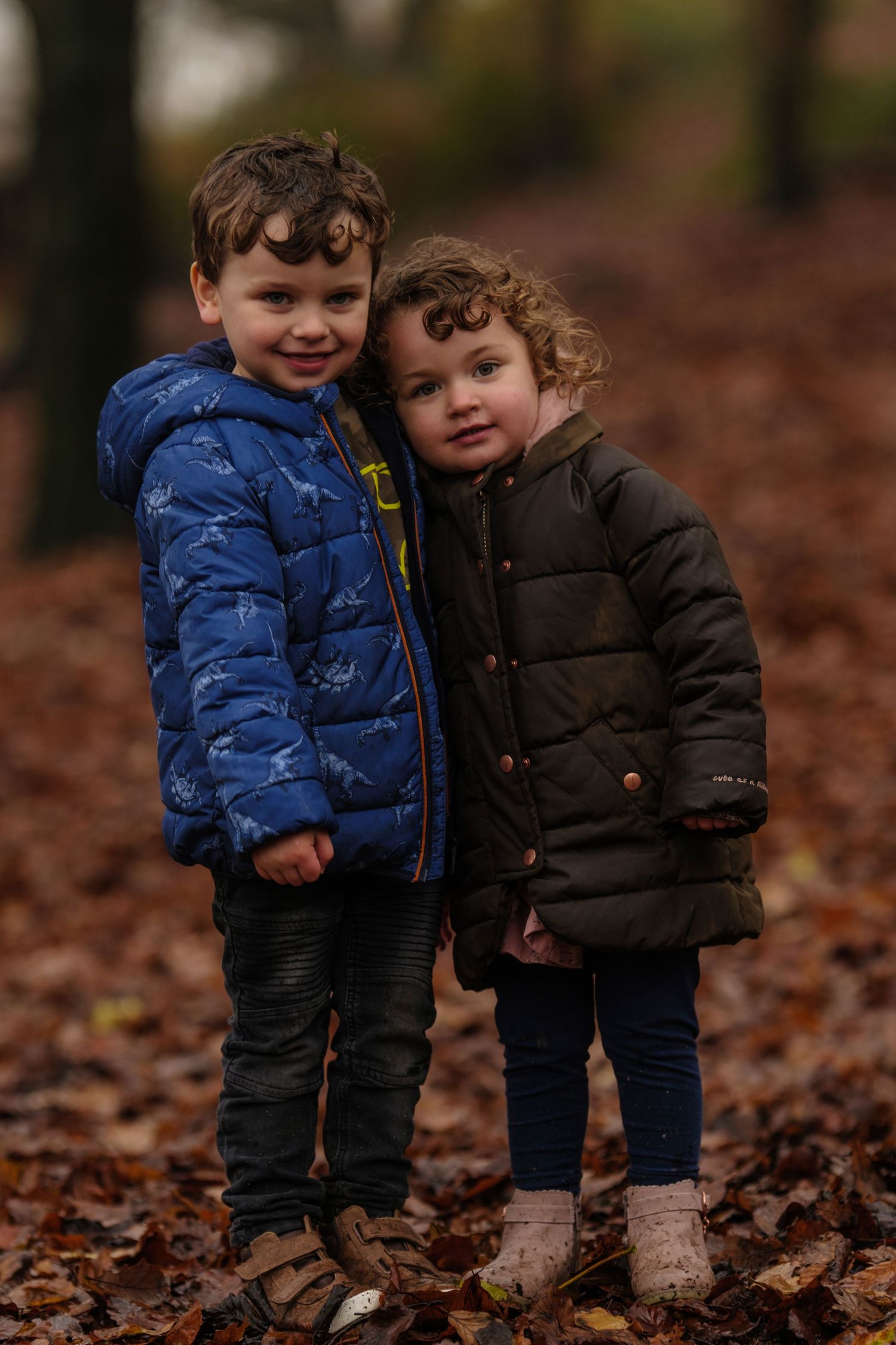 Two children in puffy winter coats stand together on fallen autumn leaves in the woods.