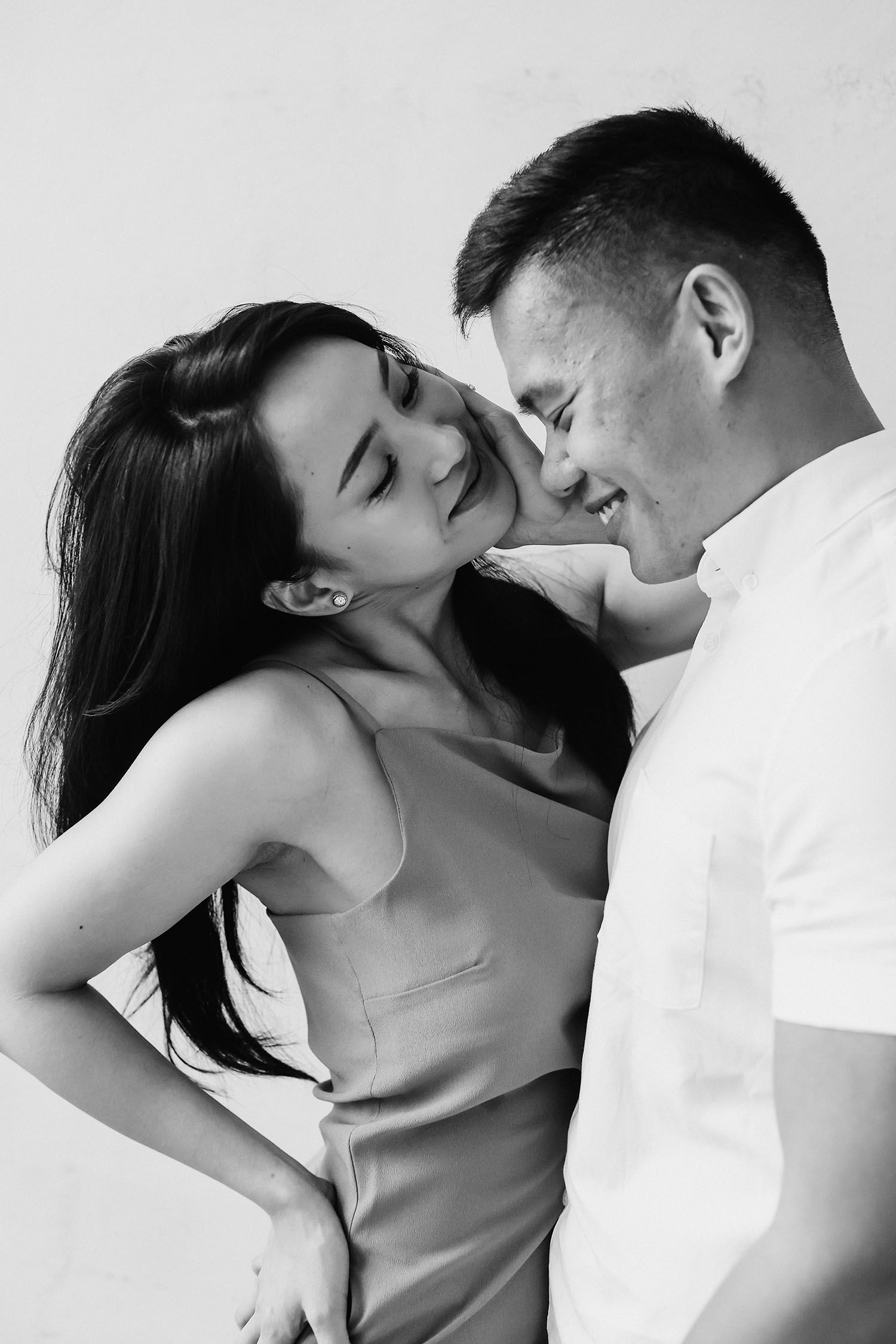 A romantic black and white portrait of a couple in an intimate moment against a plain background.