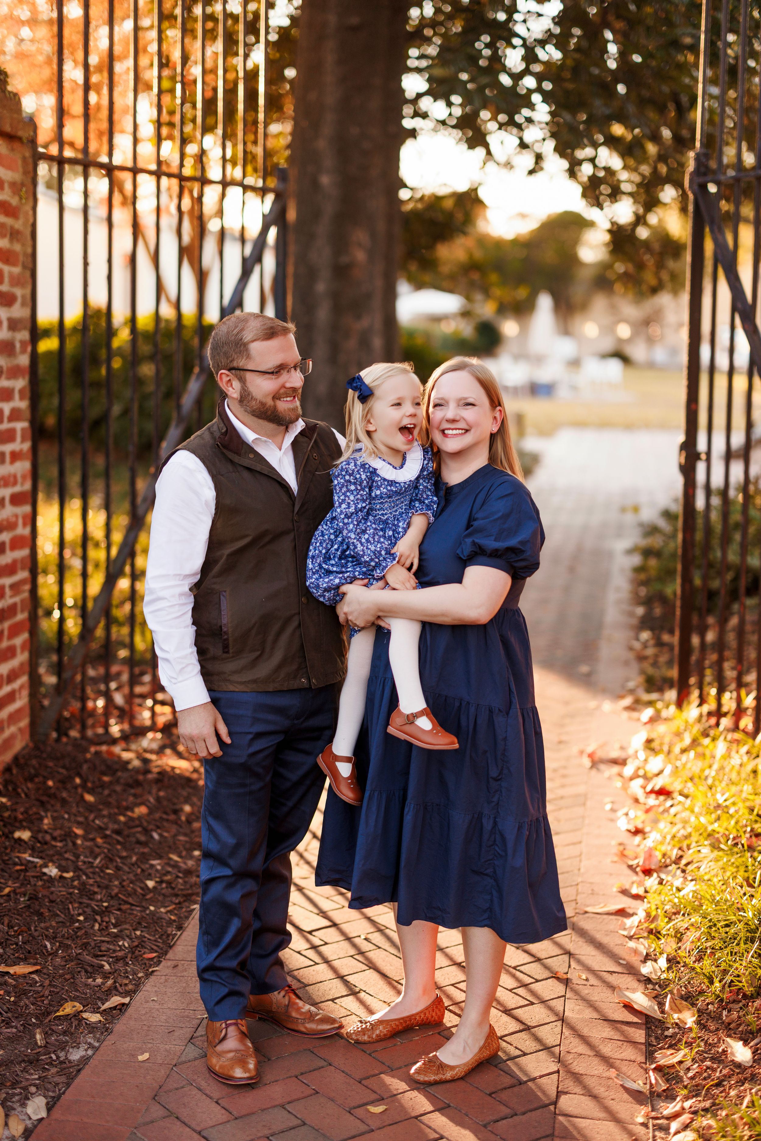 A family in coordinating navy and brown outfits poses together near a brick wall and iron gate at sunset.