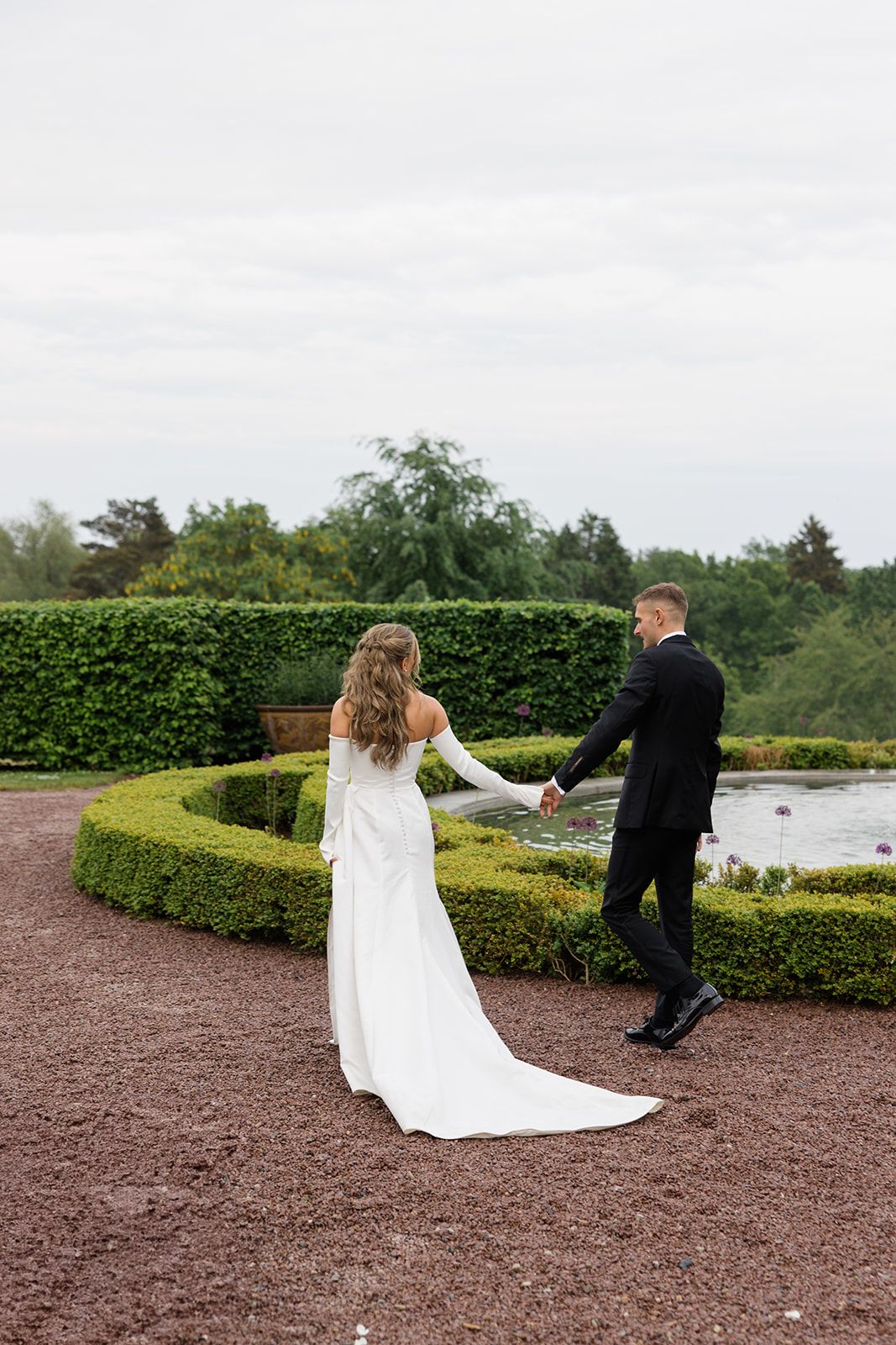 A couple in wedding attire dances playfully on a gravel path bordered by manicured hedges near a lake.