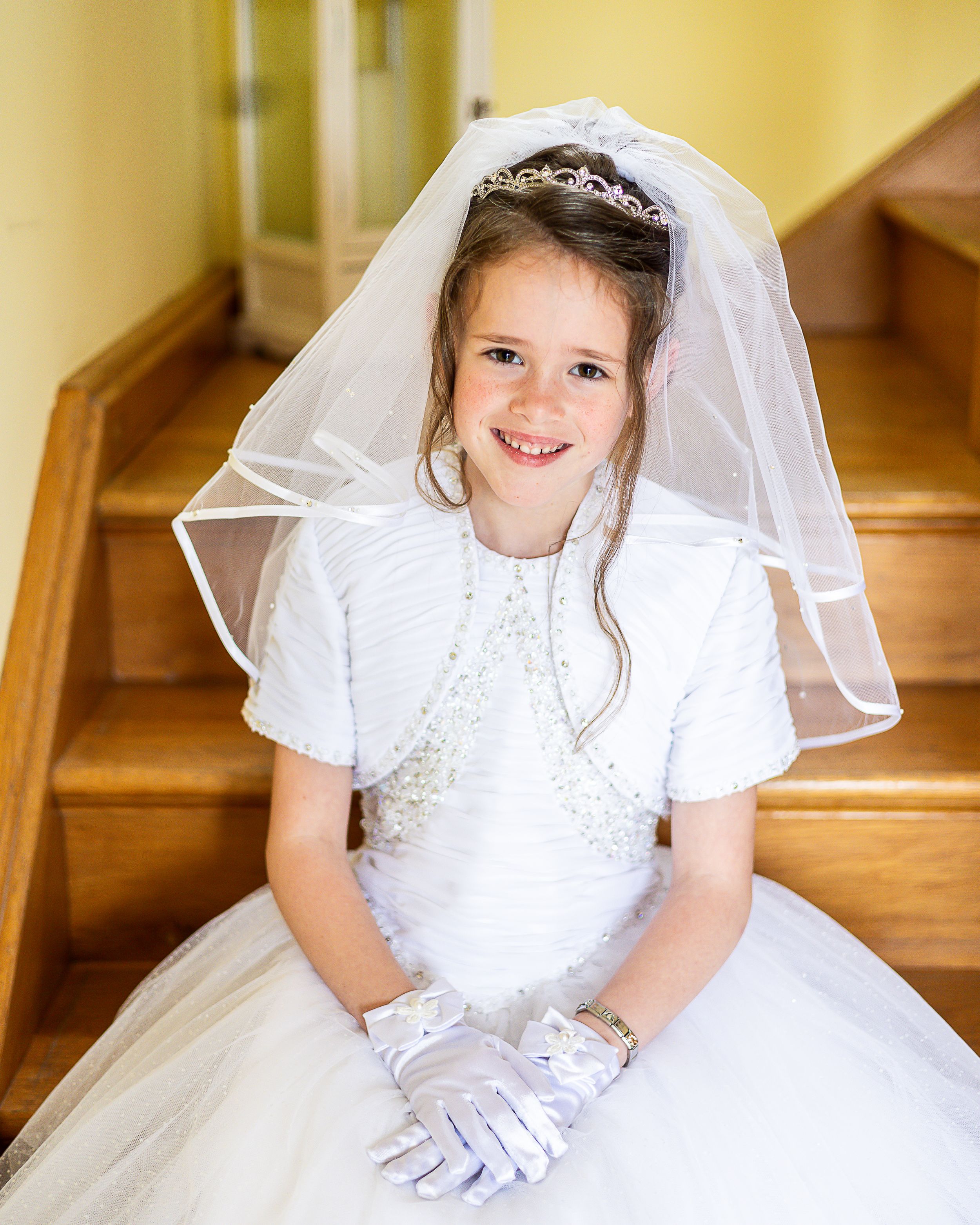 Young girl in white First Communion dress and veil poses gracefully on wooden staircase in natural light.  Newtownbutler