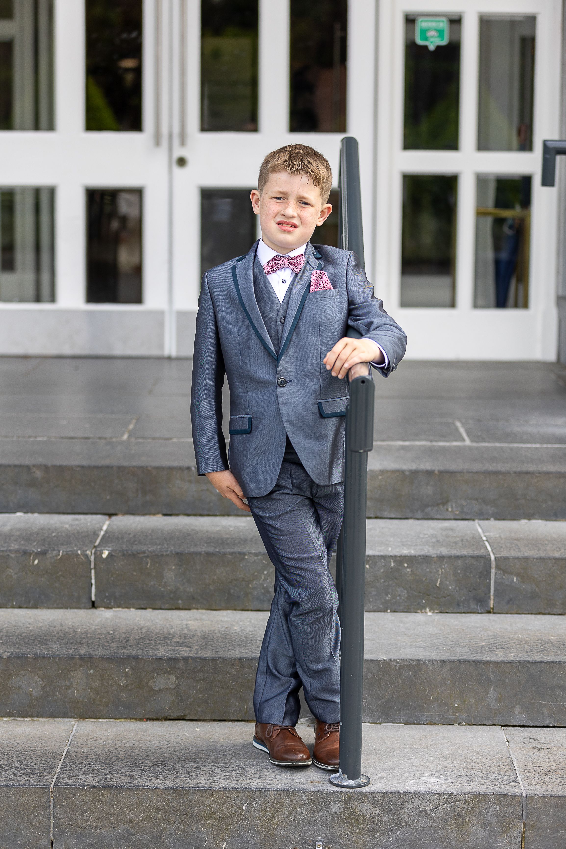 Young child in a formal grey suit with bow tie and brown shoes poses on stone steps outside glass doors.  Slieve Russell County Cavan