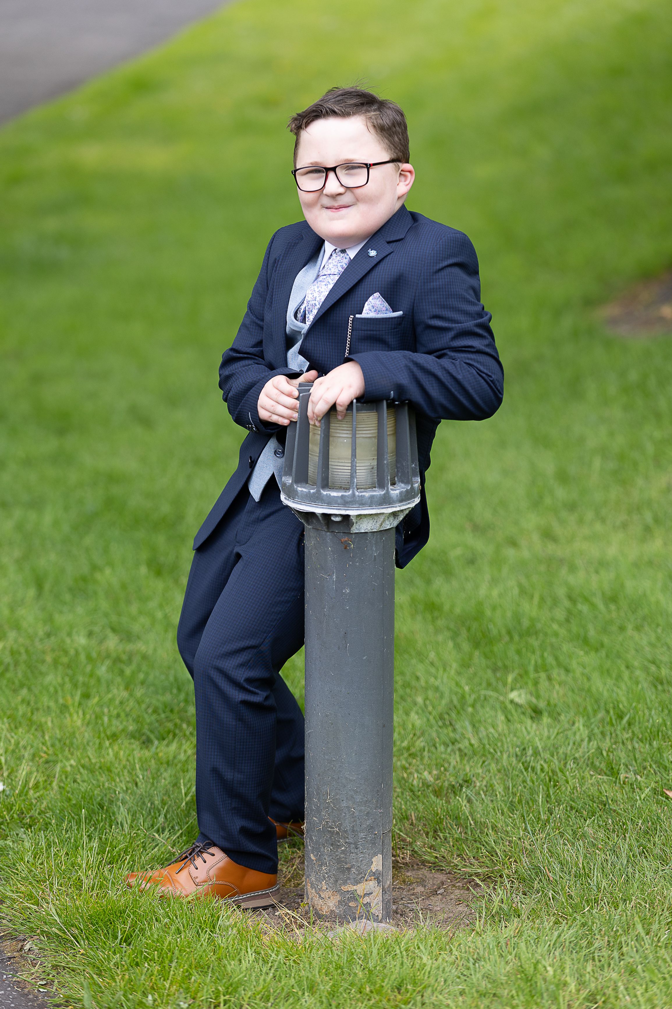 A young boy making his first communion in a navy suit sits on a metal post in a grassy outdoor setting.  Enniskillen County Fermanagh