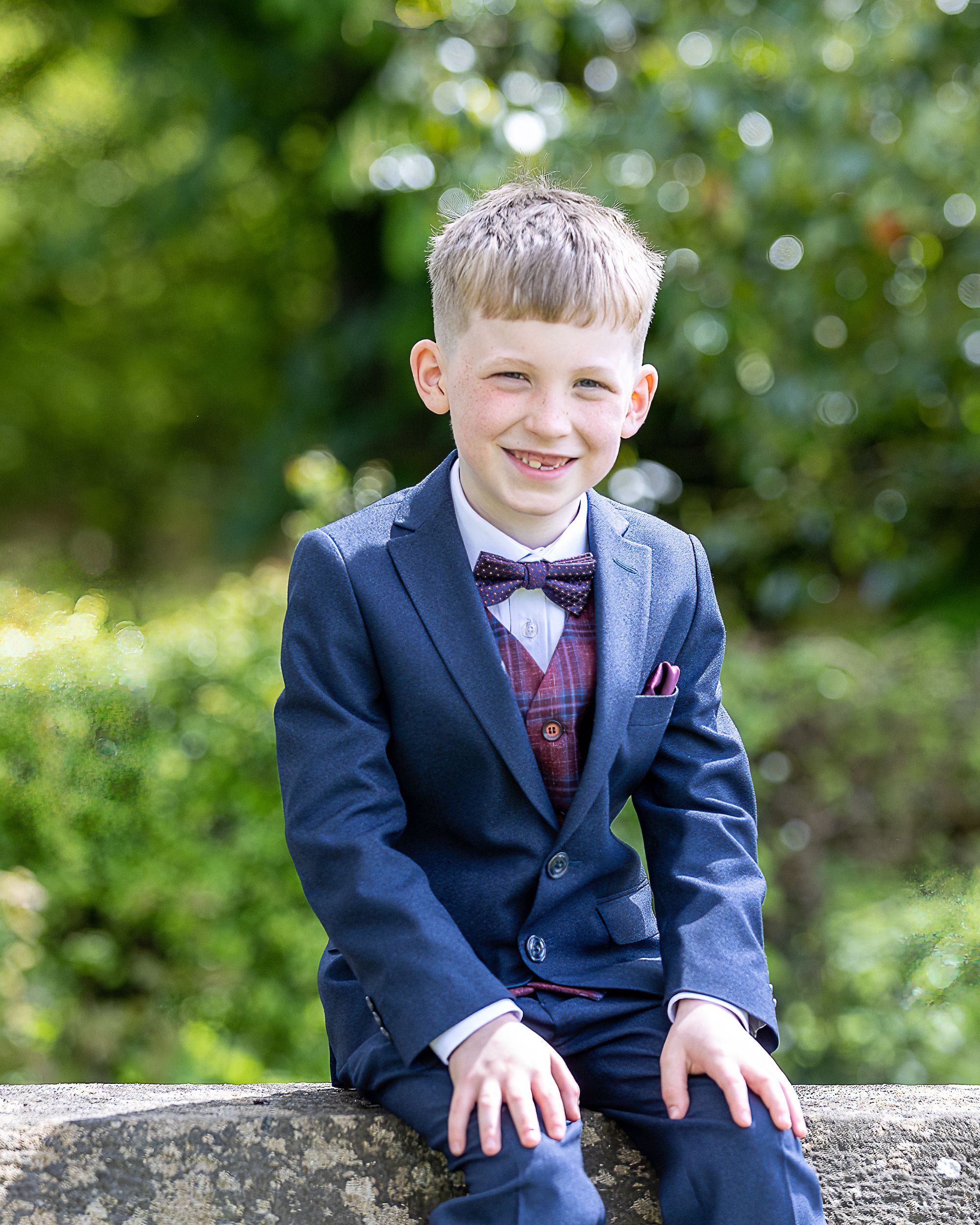 Young boy in navy suit and bow tie sits on stone wall in outdoor garden setting with natural bokeh background.  Lisnaskea Communions