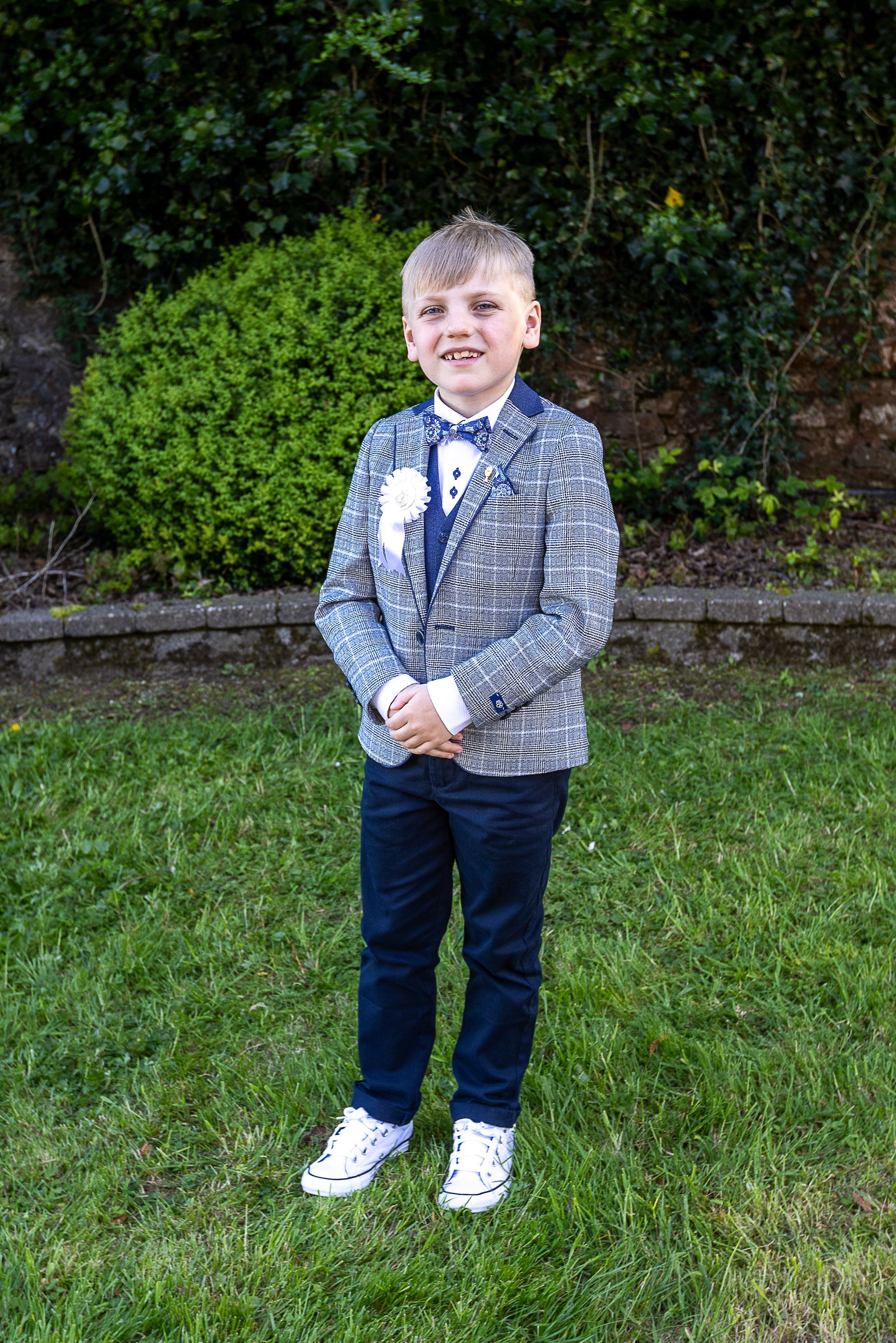 A young child wearing a plaid blazer, navy pants and white sneakers poses outdoors on green grass.  Lisbellaw