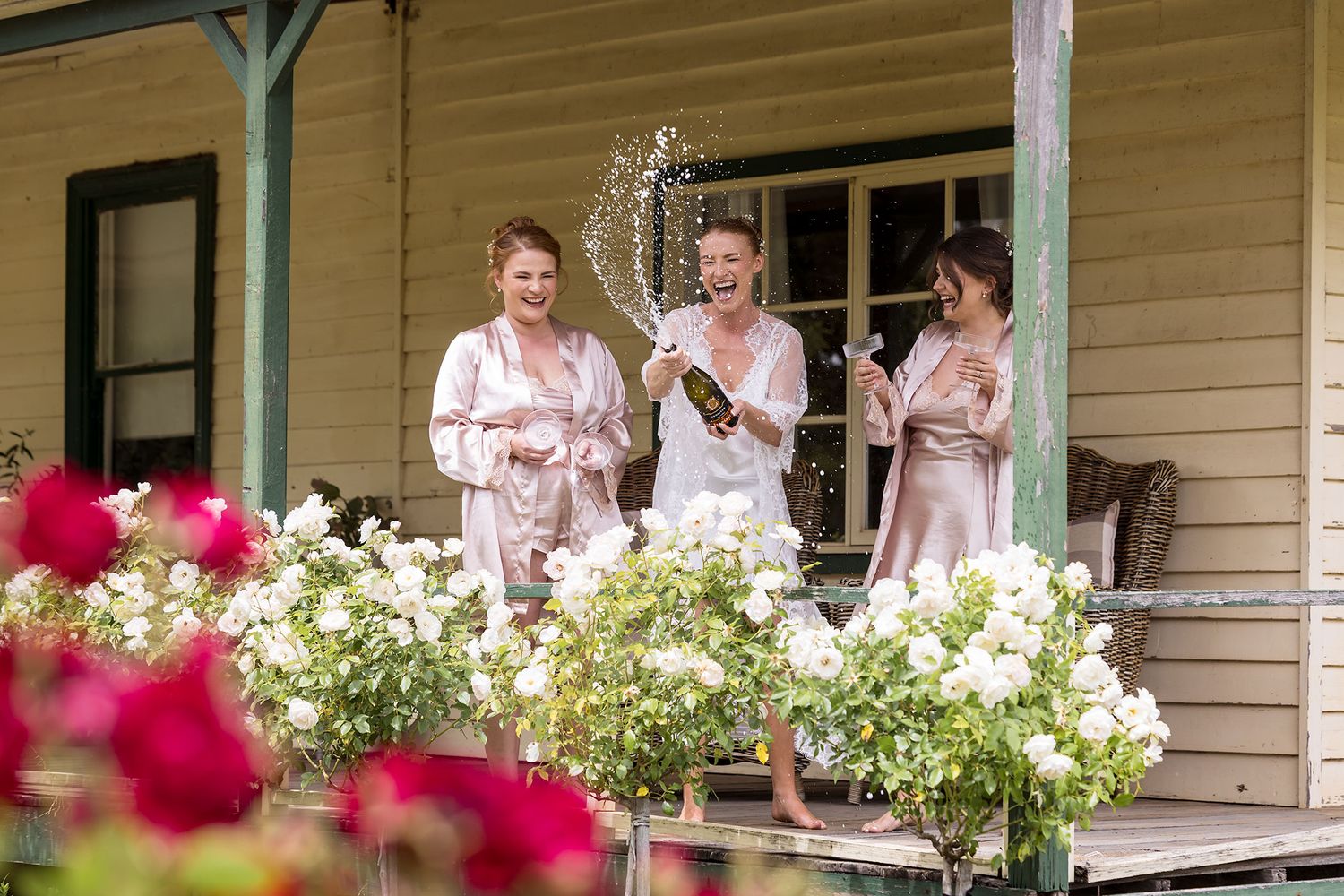 People in light-colored dresses stand on a white porch with pink and white flowers blooming in window boxes spraying champagne..