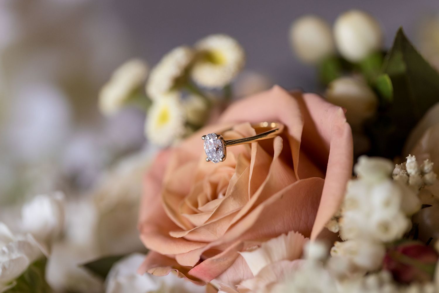 An engagement ring placed in Peach colored roses and white baby's breath flowers arranged in a delicate wedding bouquet.